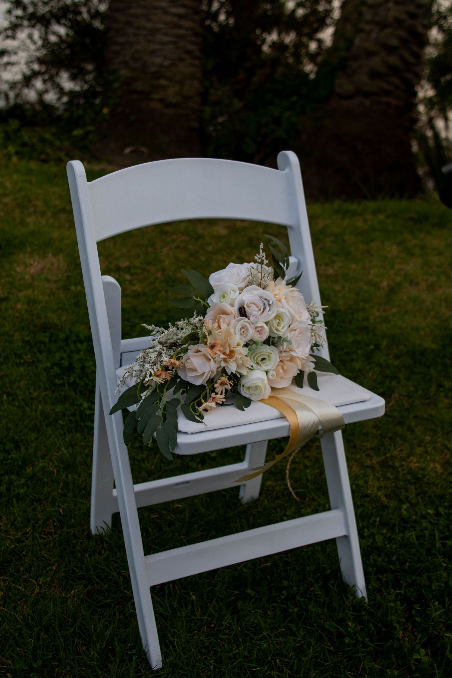 Elegant white bridal bouquet with roses and greenery resting on a white folding chair outdoors.