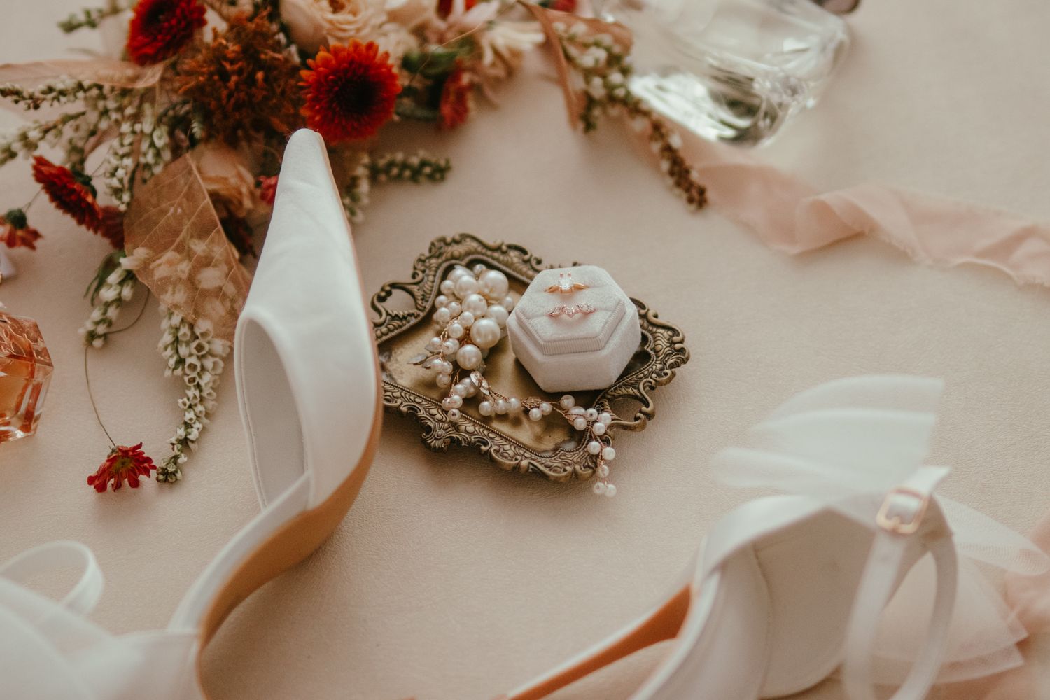 Elegant bridal flat lay with white heels, vintage ring box, pearl details, and red floral arrangement on blush background.