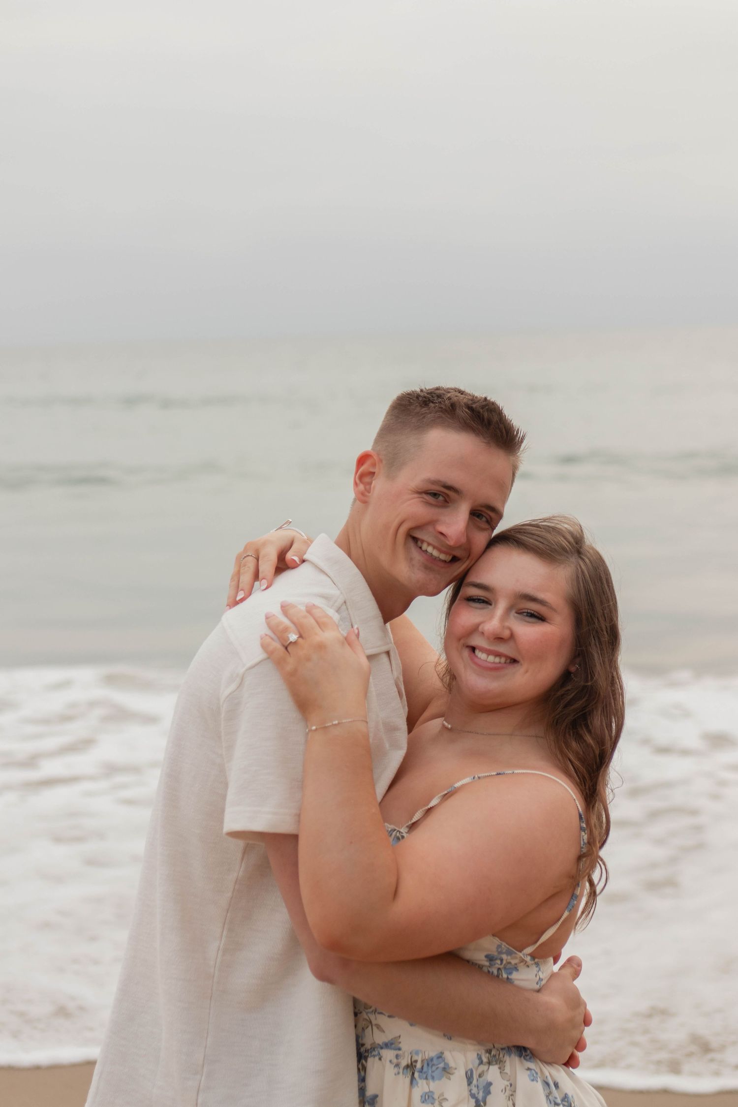 Happy smiling couple hugging on a beach with ocean waves in the background on a cloudy day.