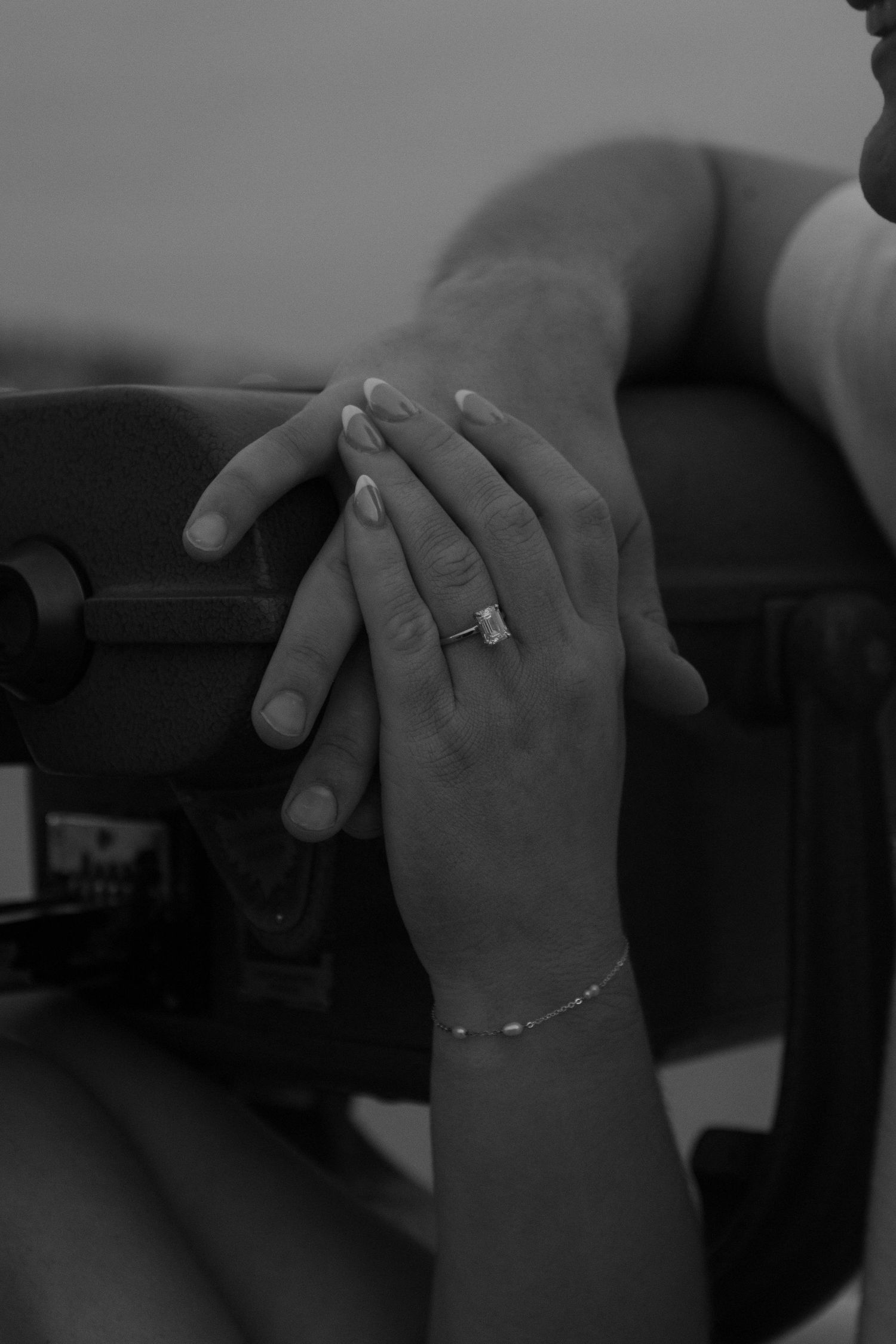 Close-up black and white photo of hands with an emerald cut engagement ring and delicate bracelet.