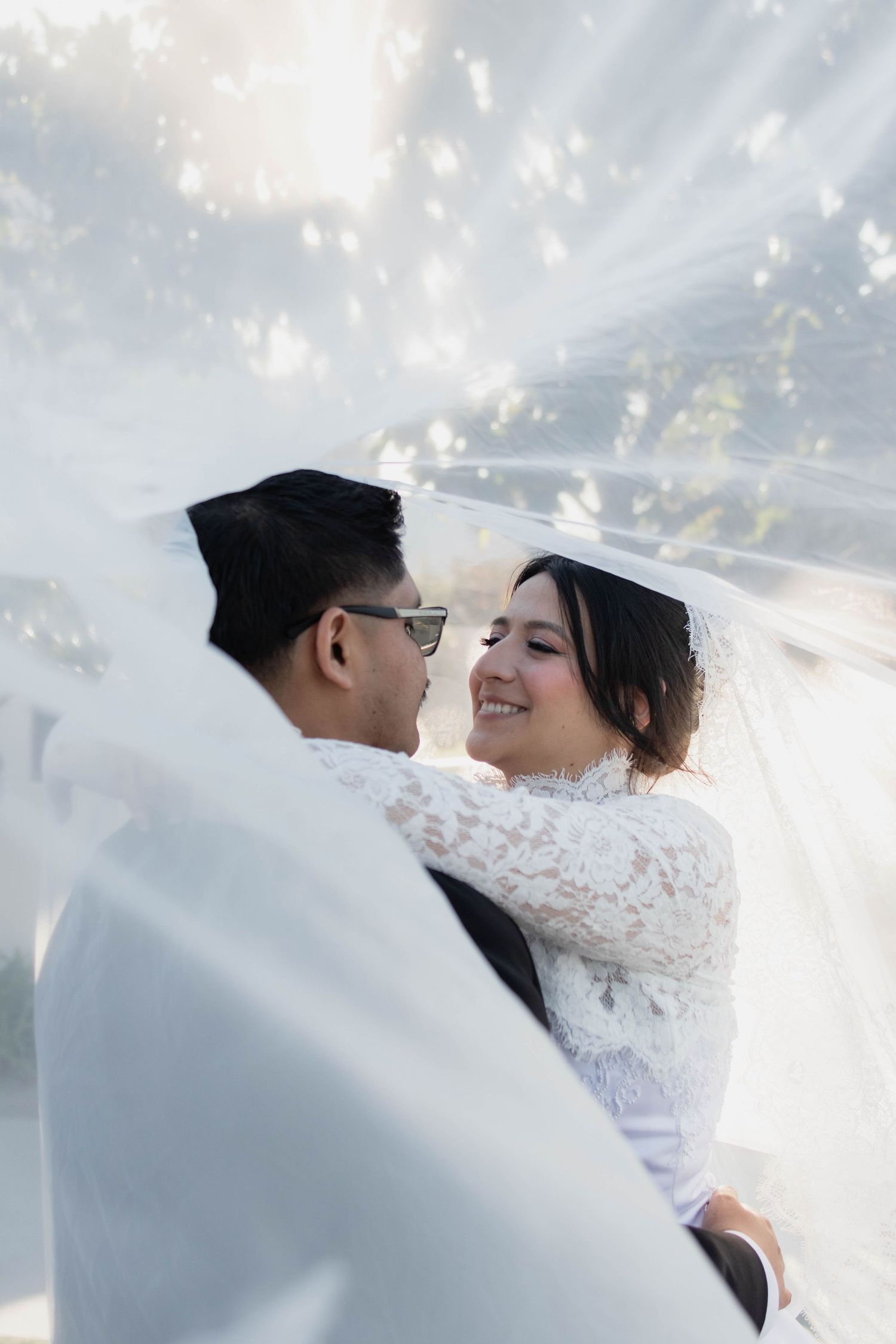 Smiling bride in lace gown gazes at groom under billowing white veil in dreamy sunlit setting.