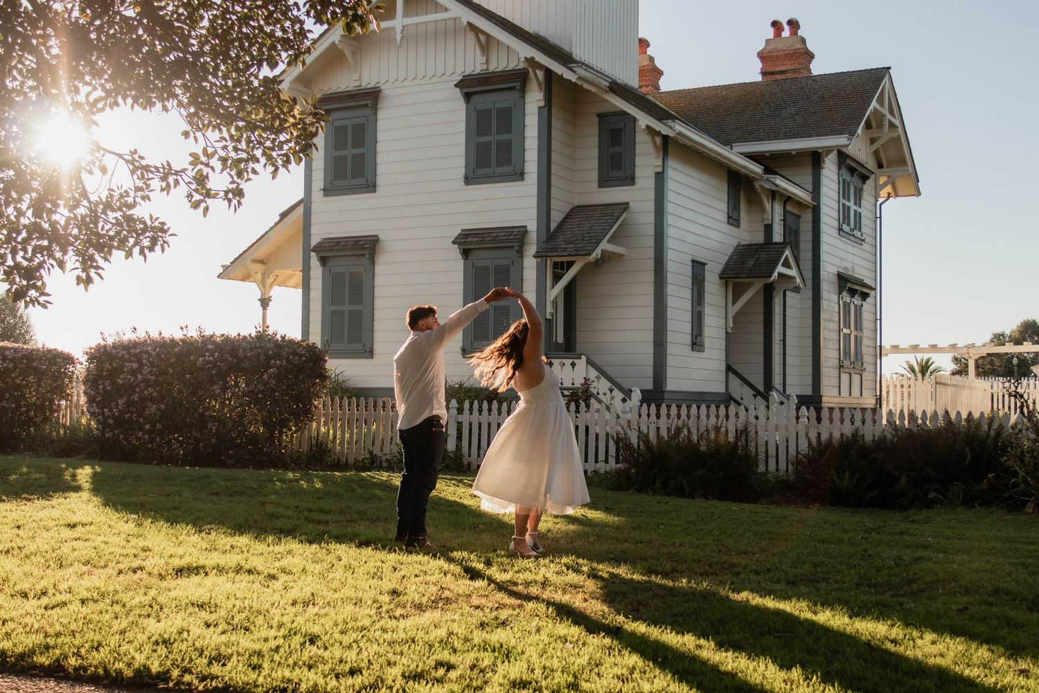 Couple dances joyfully on green lawn before charming white lighthouse at golden hour sunset.