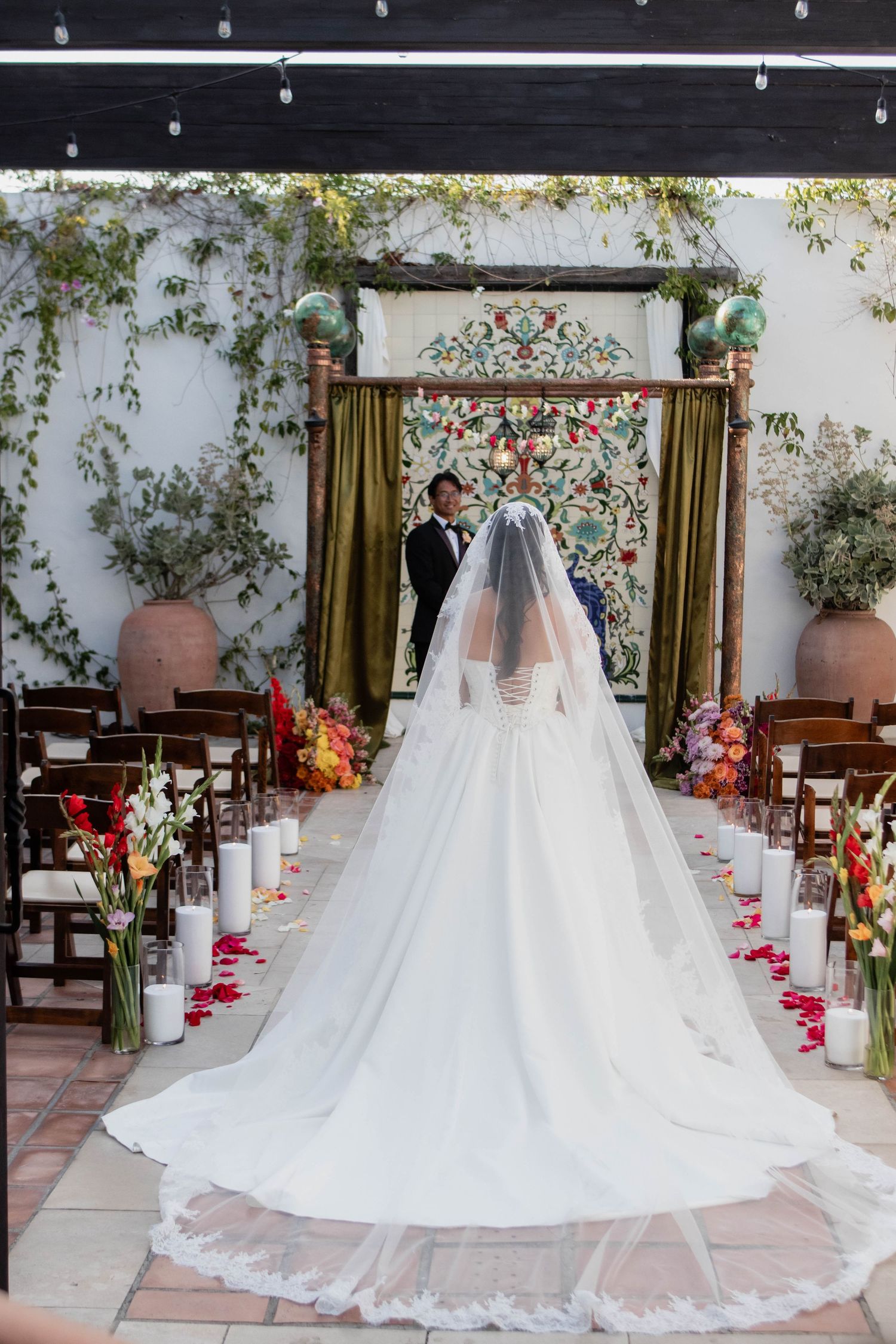 Bride in a long white gown and veil walks toward groom at an outdoor wedding ceremony with floral decor.