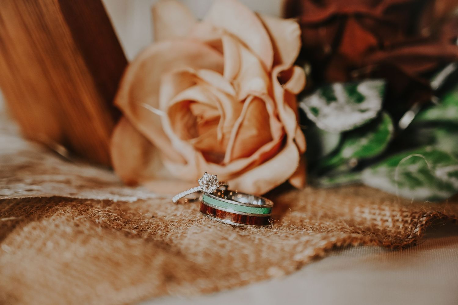 Wedding rings with diamond halo setting displayed on burlap beside a peach rose and greenery.