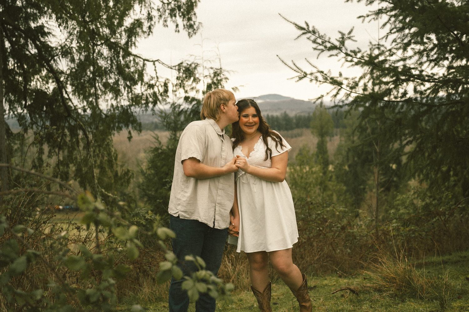 Couple sharing a romantic moment among evergreen trees with misty mountain backdrop in an outdoor engagement photo session.