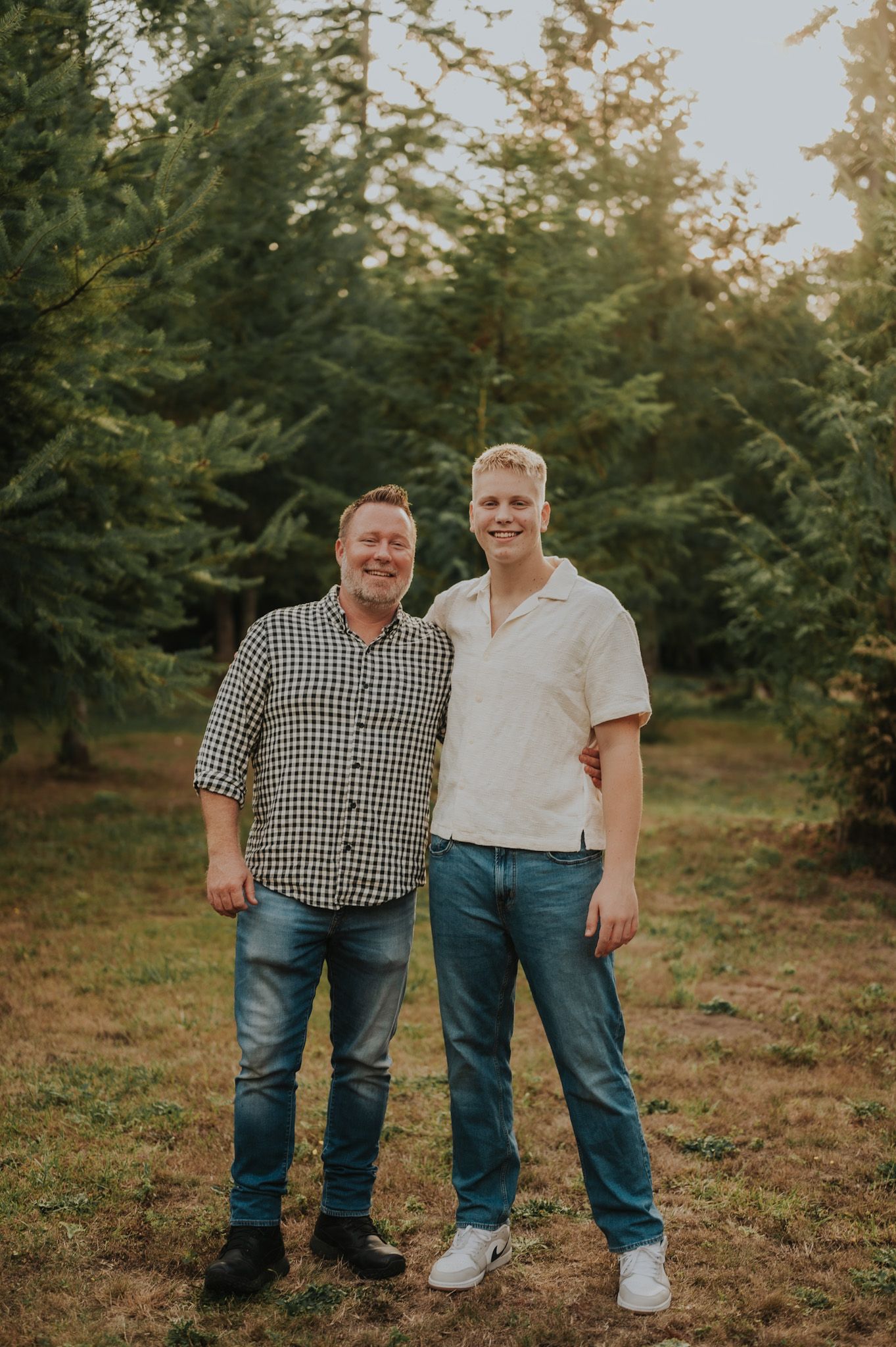 Two smiling men stand together outdoors among green trees during golden hour in a casual portrait photo.