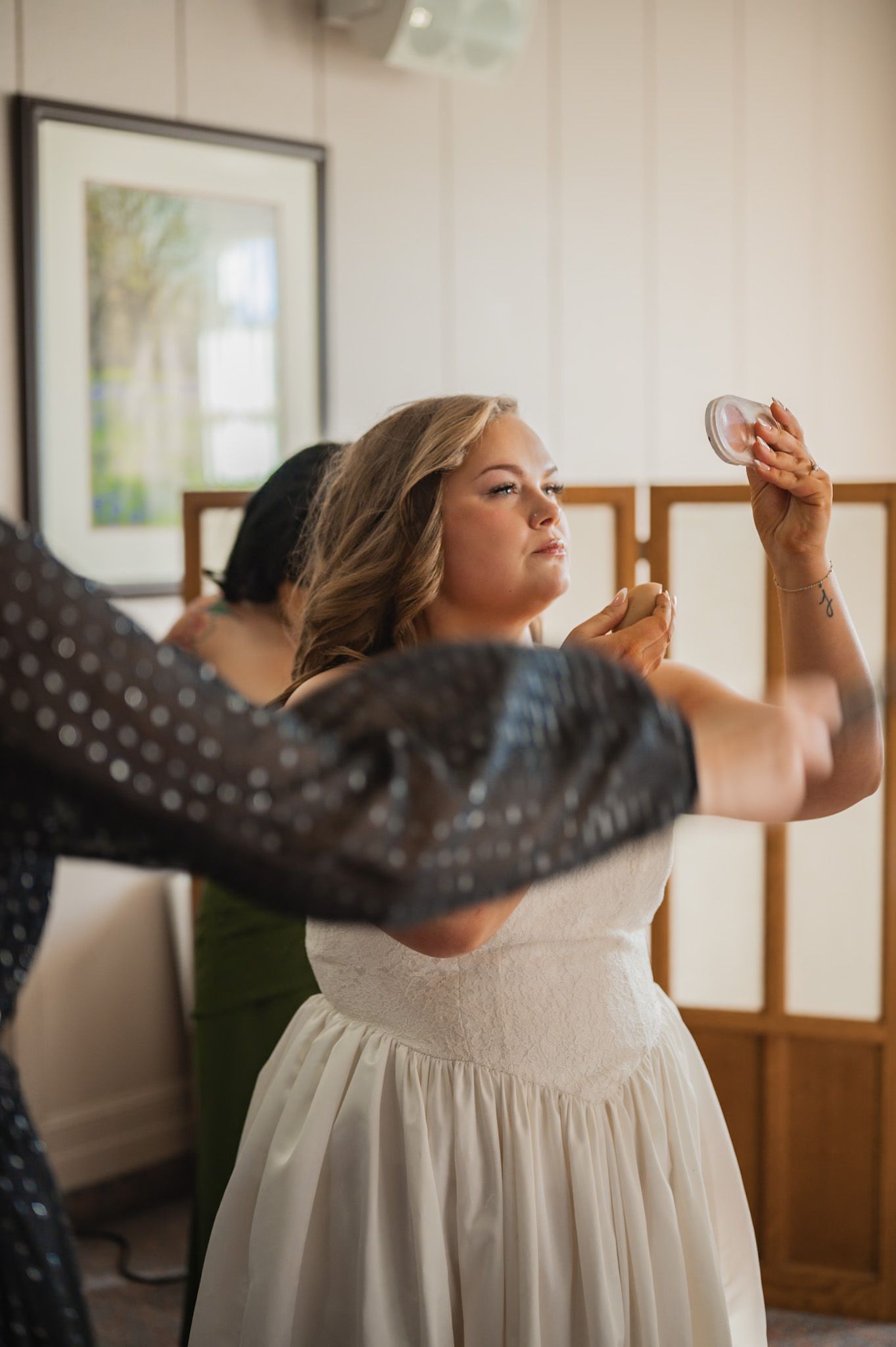 Bride in white strapless gown holds a compact mirror while being helped by a bridesmaid in a polka dot dress.