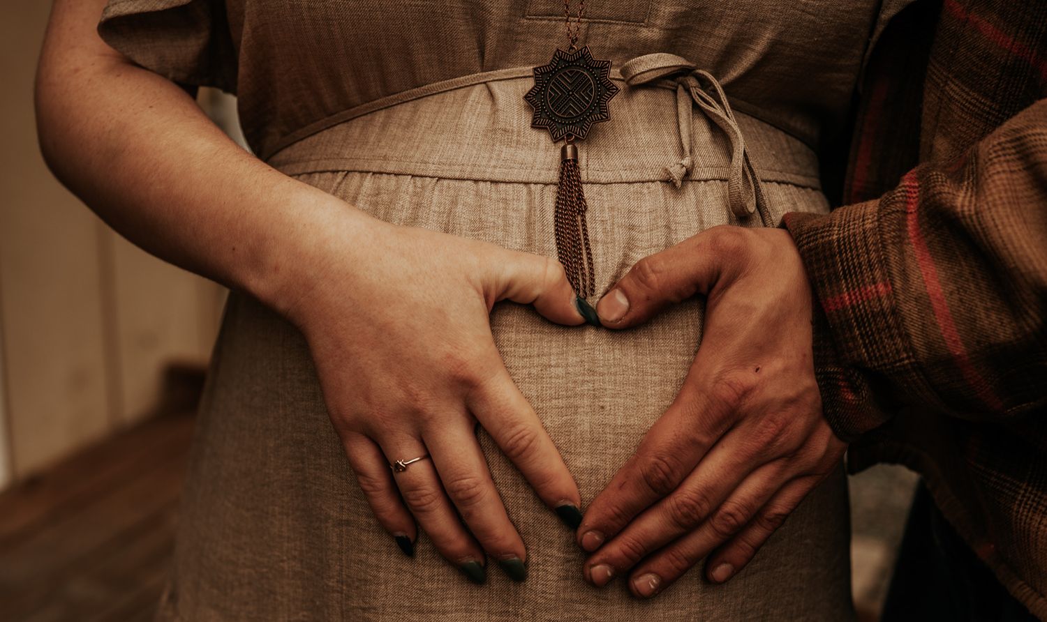 Couple forming a heart shape with their hands on a pregnant woman's belly, wearing casual earthy toned clothing.
