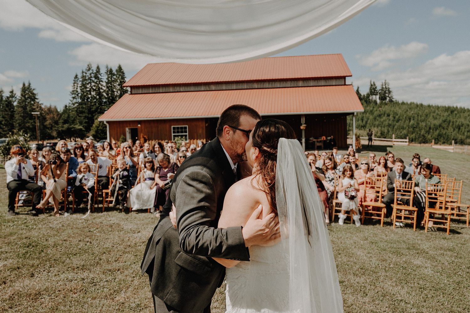 Bride and groom share first kiss at outdoor barn wedding ceremony with guests seated on wooden chairs.