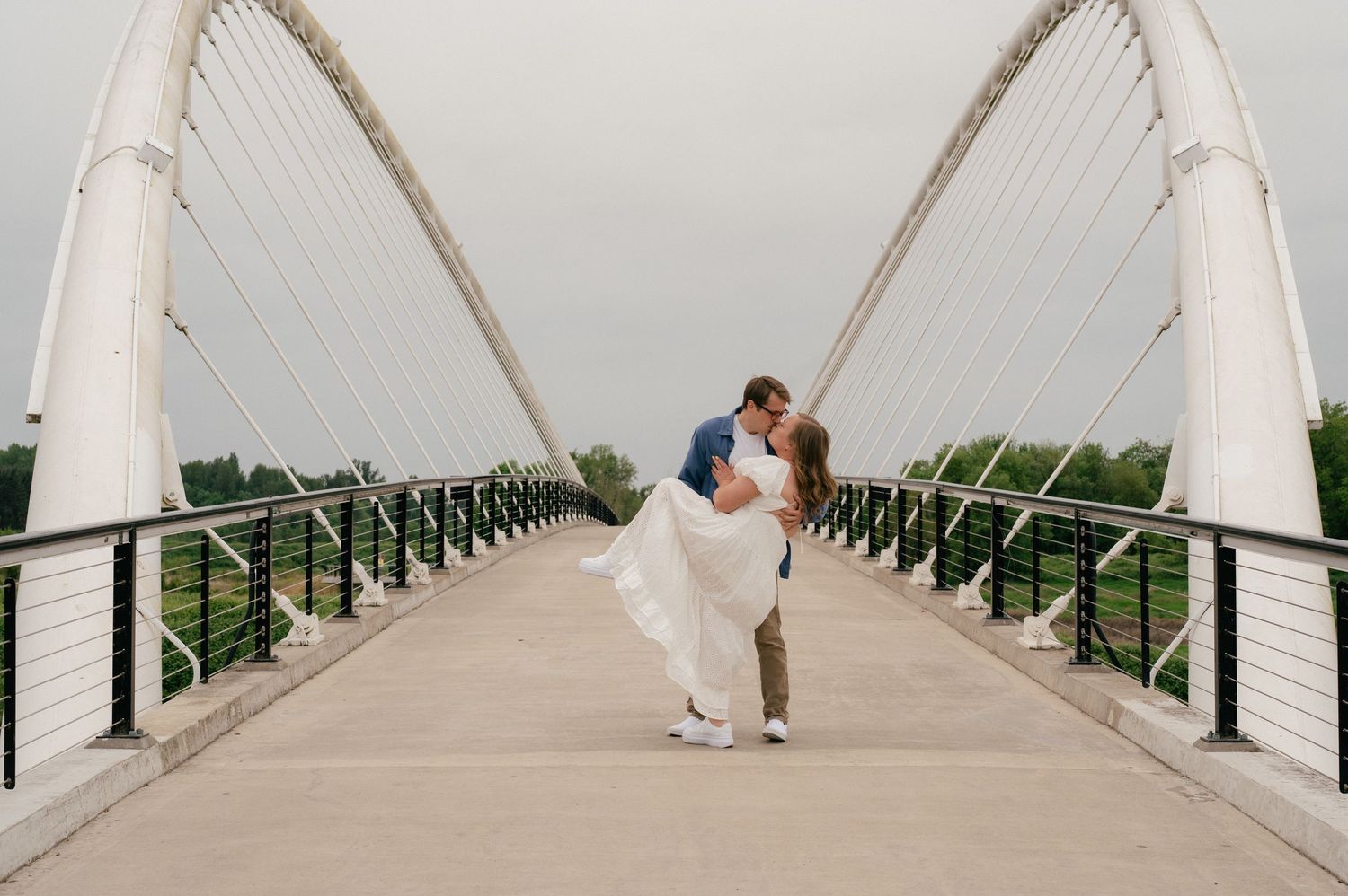 Bride and groom share romantic dip kiss on white cable suspension bridge, wedding dress flowing, lush green trees background.