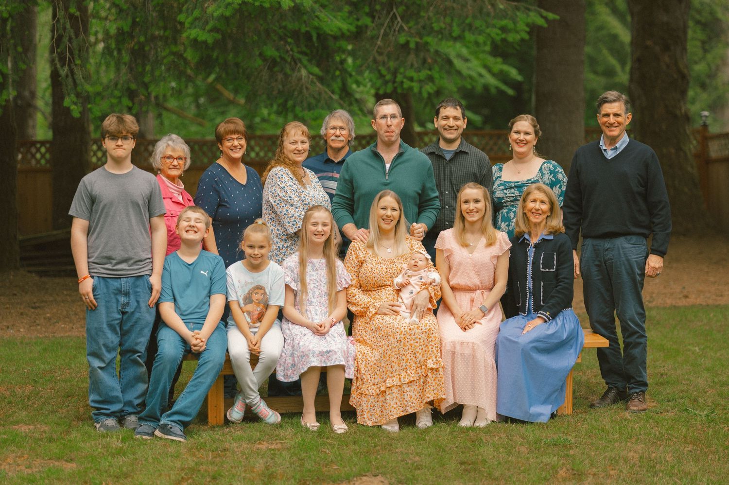 Large family group portrait outdoors among tall pine trees, featuring adults and children posing together on a bench.