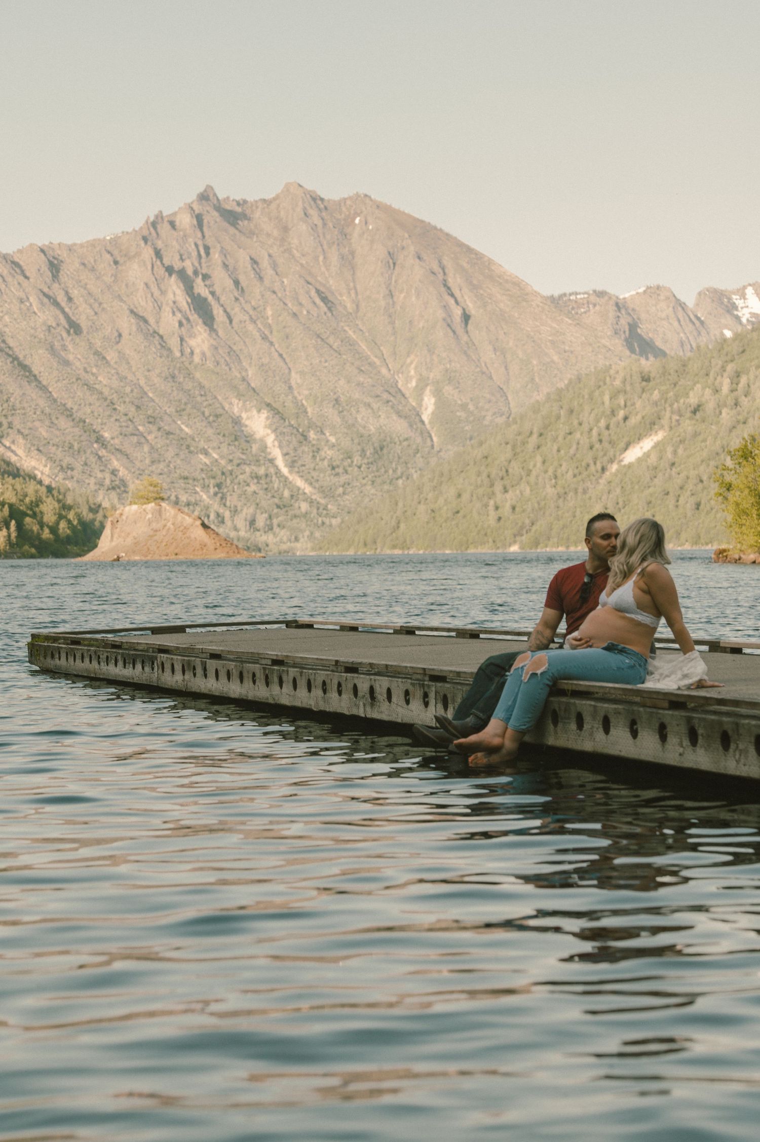 Couple sitting on a floating dock sharing a romantic moment with mountain lake views in the background.