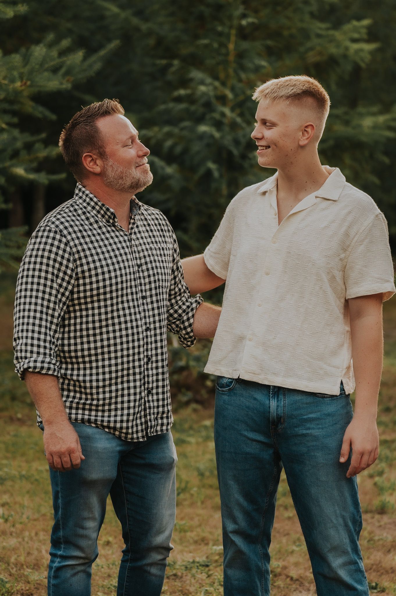 Two men smiling at each other outdoors near evergreen trees, one in plaid shirt and one in cream top.