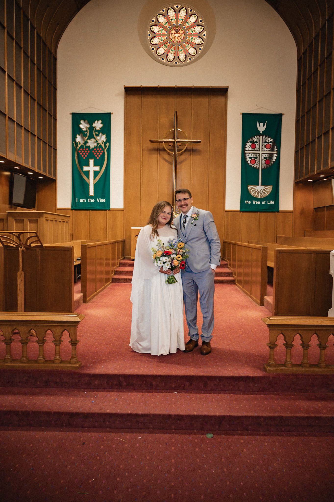 Bride and groom posing together at church altar with cross, stained glass window, and green banners on wedding day.