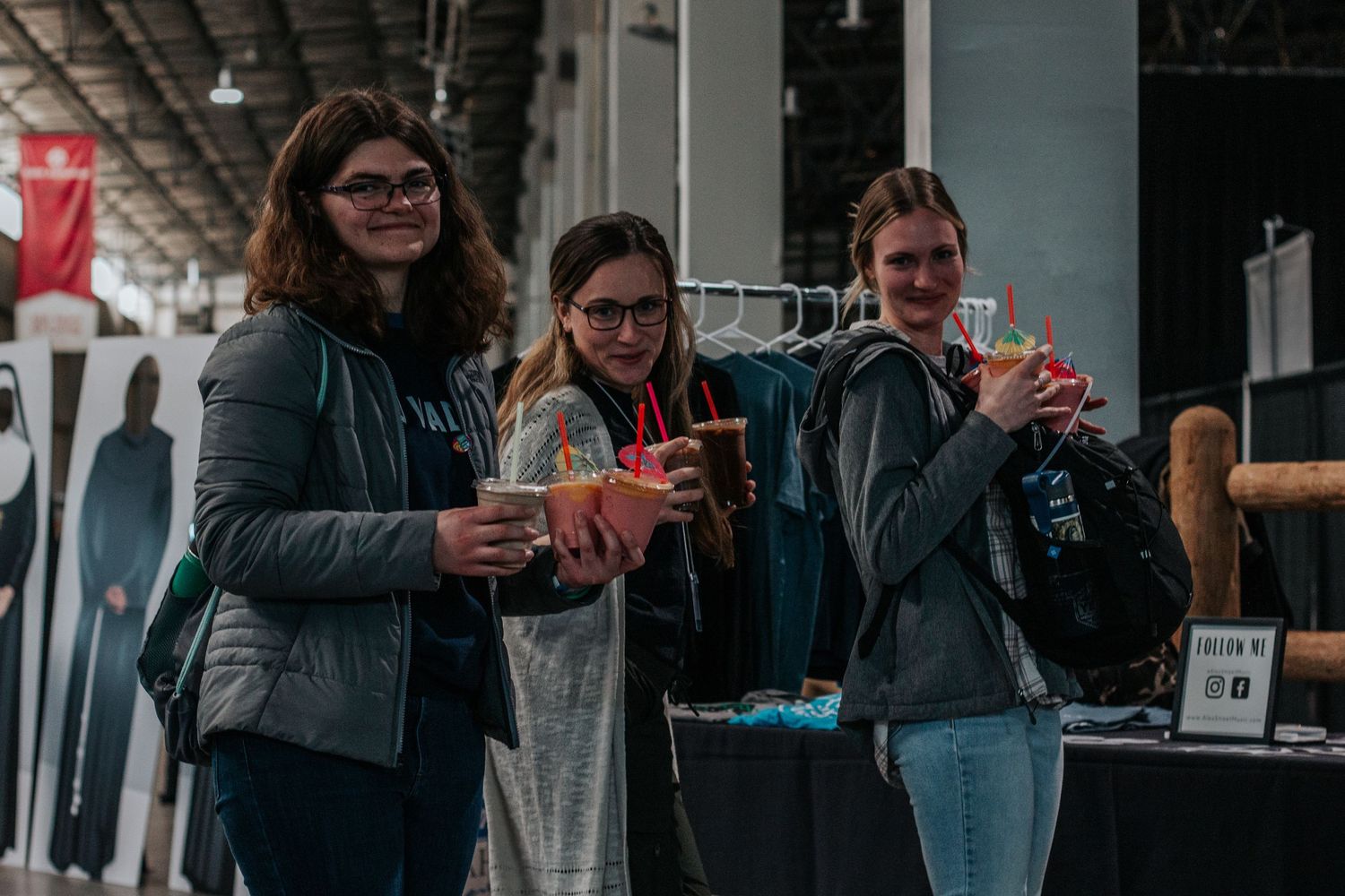 Three smiling teen girls hold cups of treats while browsing vendor tables at a large indoor youth conference.