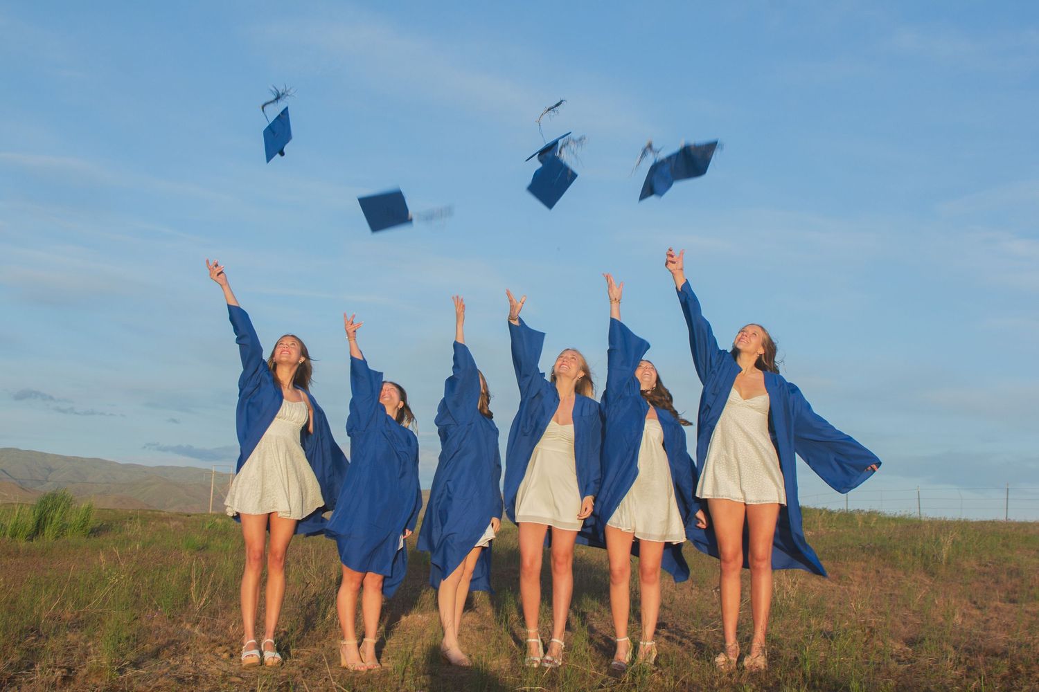 Six graduates in blue gowns toss their caps into the air in a grassy field beneath a blue sky with clouds.