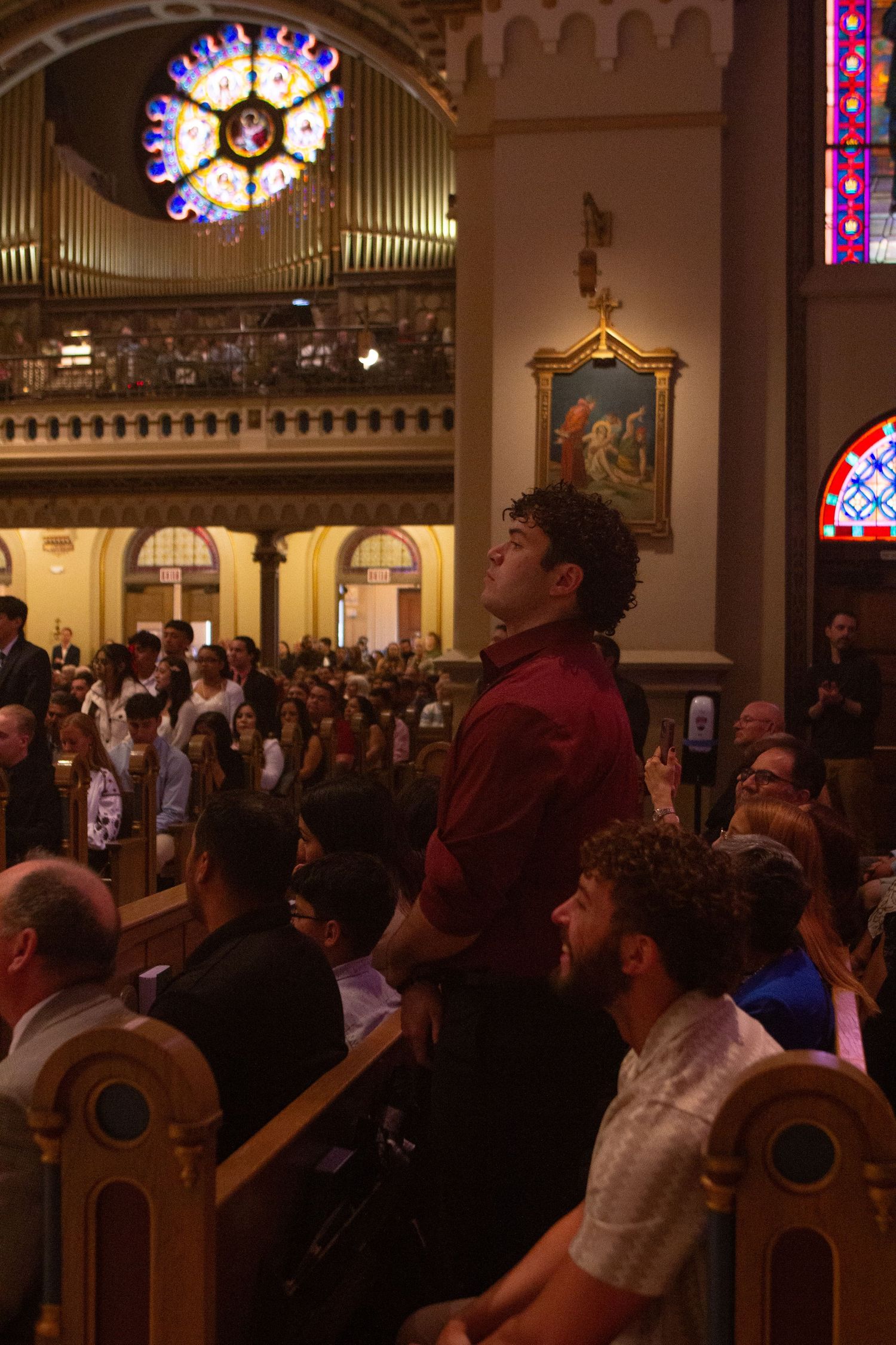 Parishioners fill the pews of an ornate Catholic church as a young man stands in the aisle gazing upward beneath a rose window.