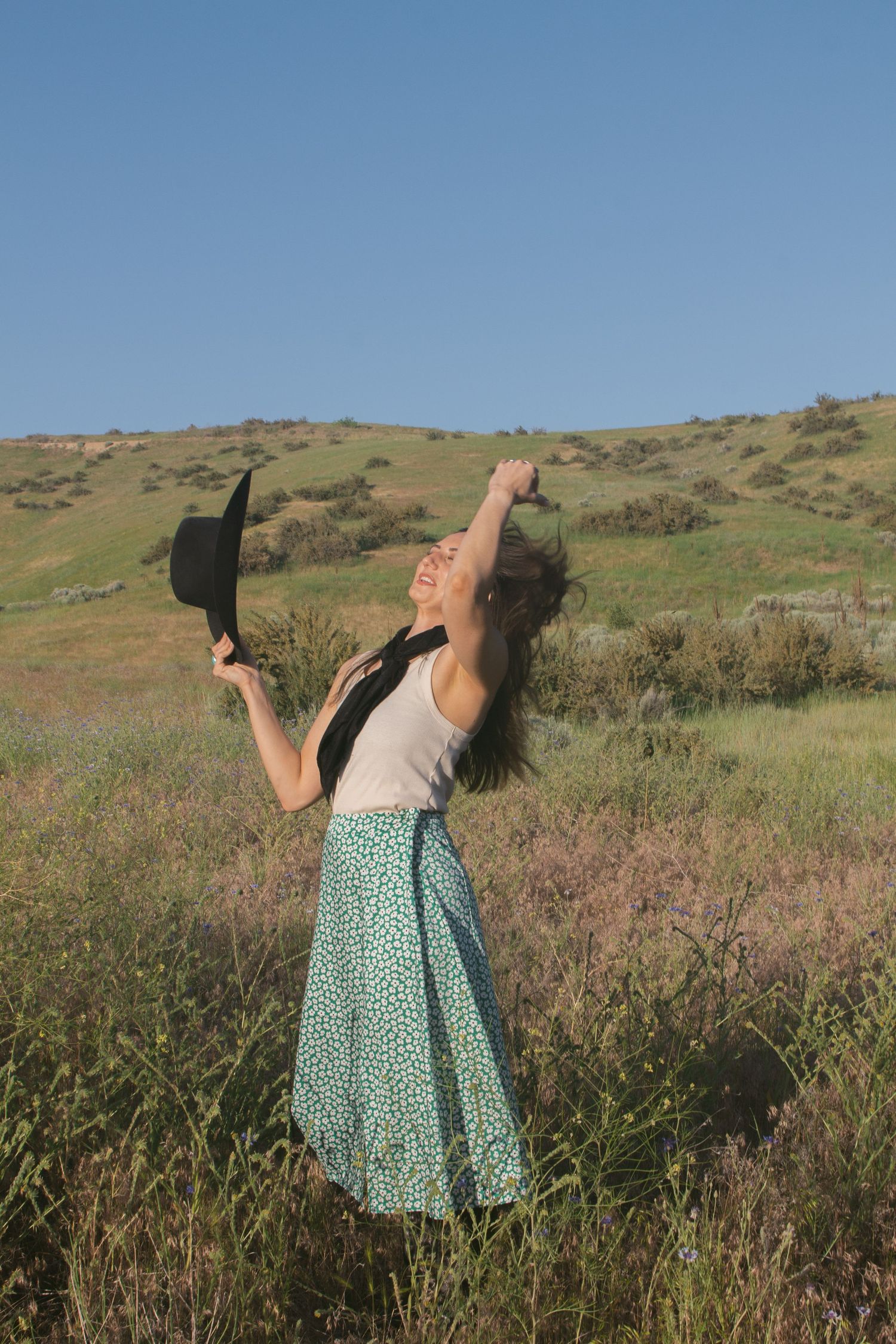 Woman in a green floral skirt tilts her head back with arms raised, holding a black hat in a sunny wildflower meadow.