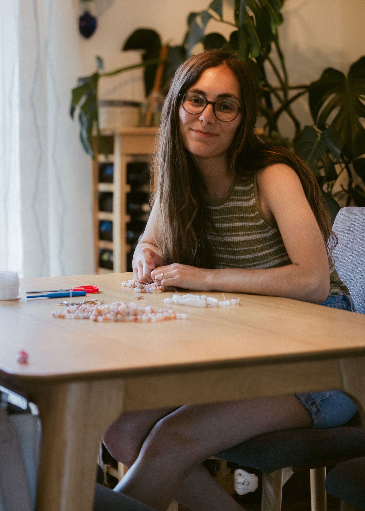 Smiling woman with glasses sits at a wooden table stringing colorful beads for a handmade jewelry project.