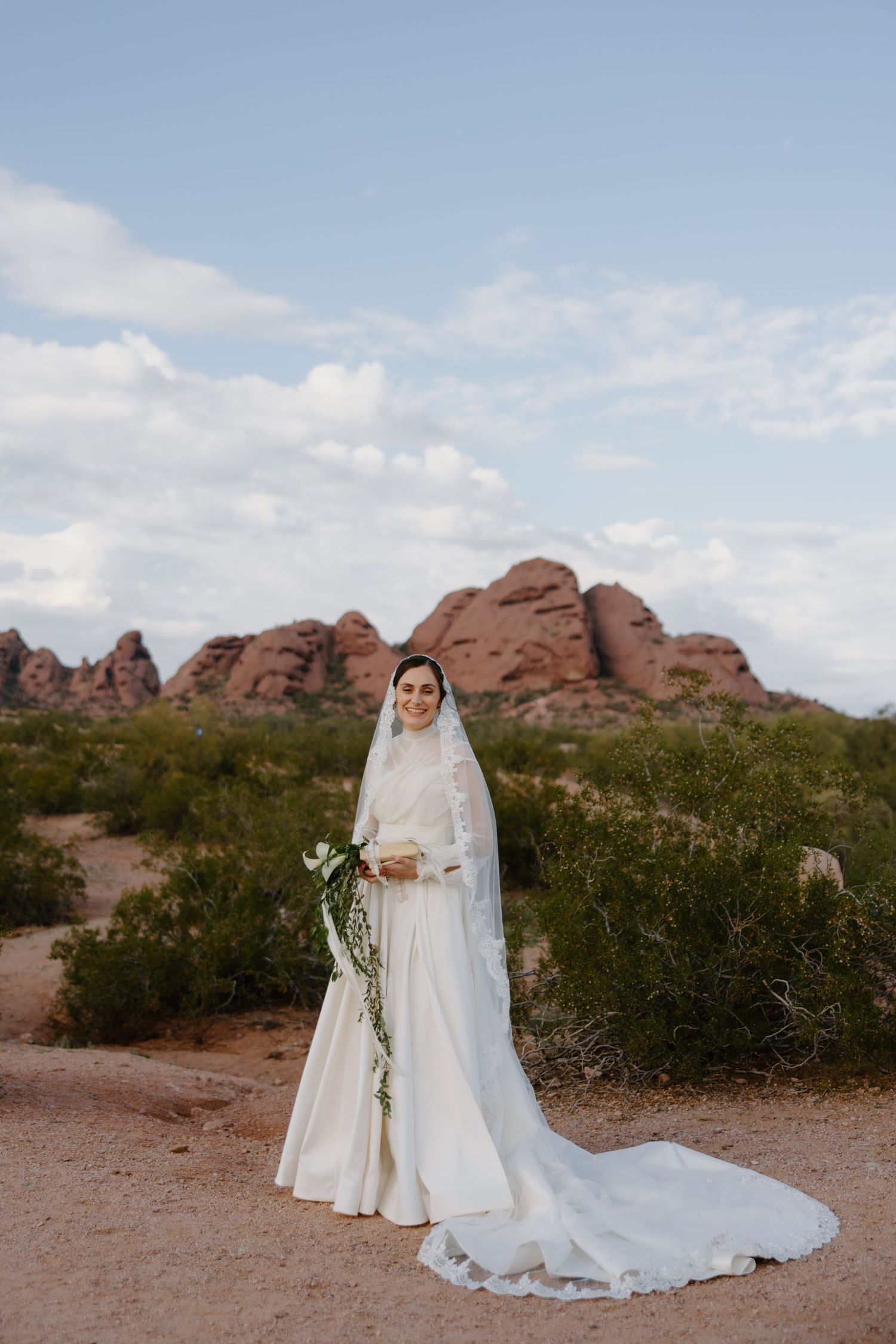 Bride in a white gown and long veil holds a green bouquet in a desert landscape with red rock formations behind her.