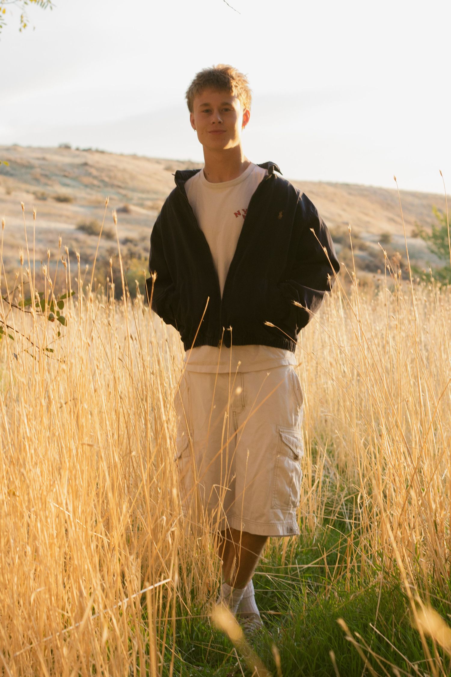 Young man in a dark hoodie and khaki shorts stands among tall golden grass in warm evening light.