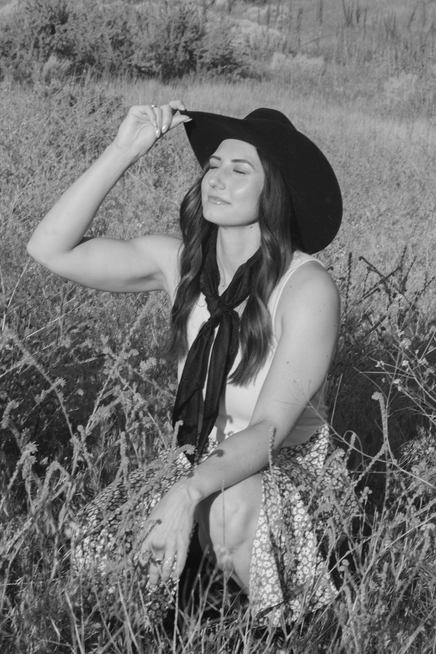 A black and white portrait of a woman in a cowboy hat and neck scarf sitting in tall grass with her eyes closed and hand on her hat.
