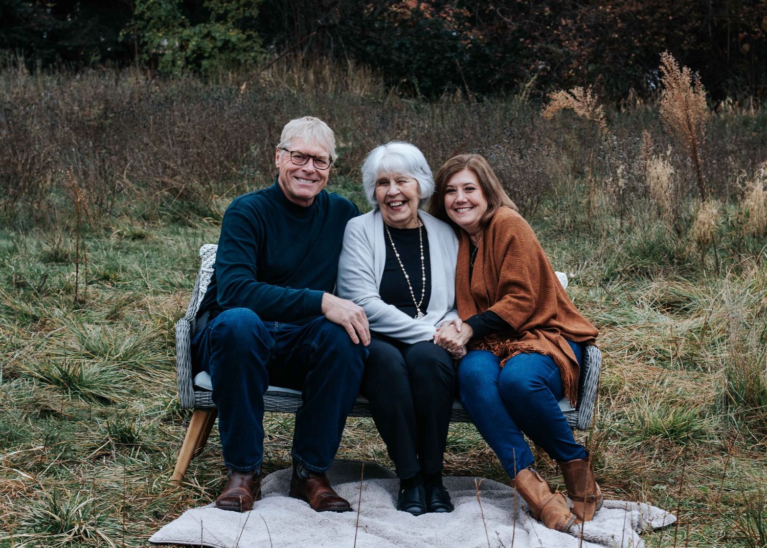An older couple and a young woman sit together on a bench in a grassy field, smiling in warm autumn family portrait.