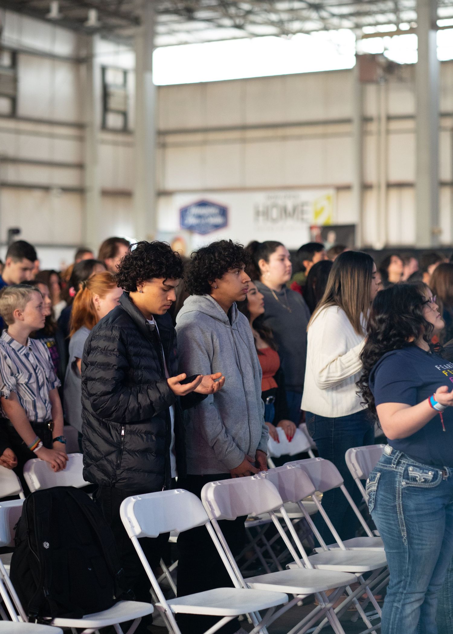 Teens and young adults stand among rows of white folding chairs in a large hall during a faith-centered youth event.