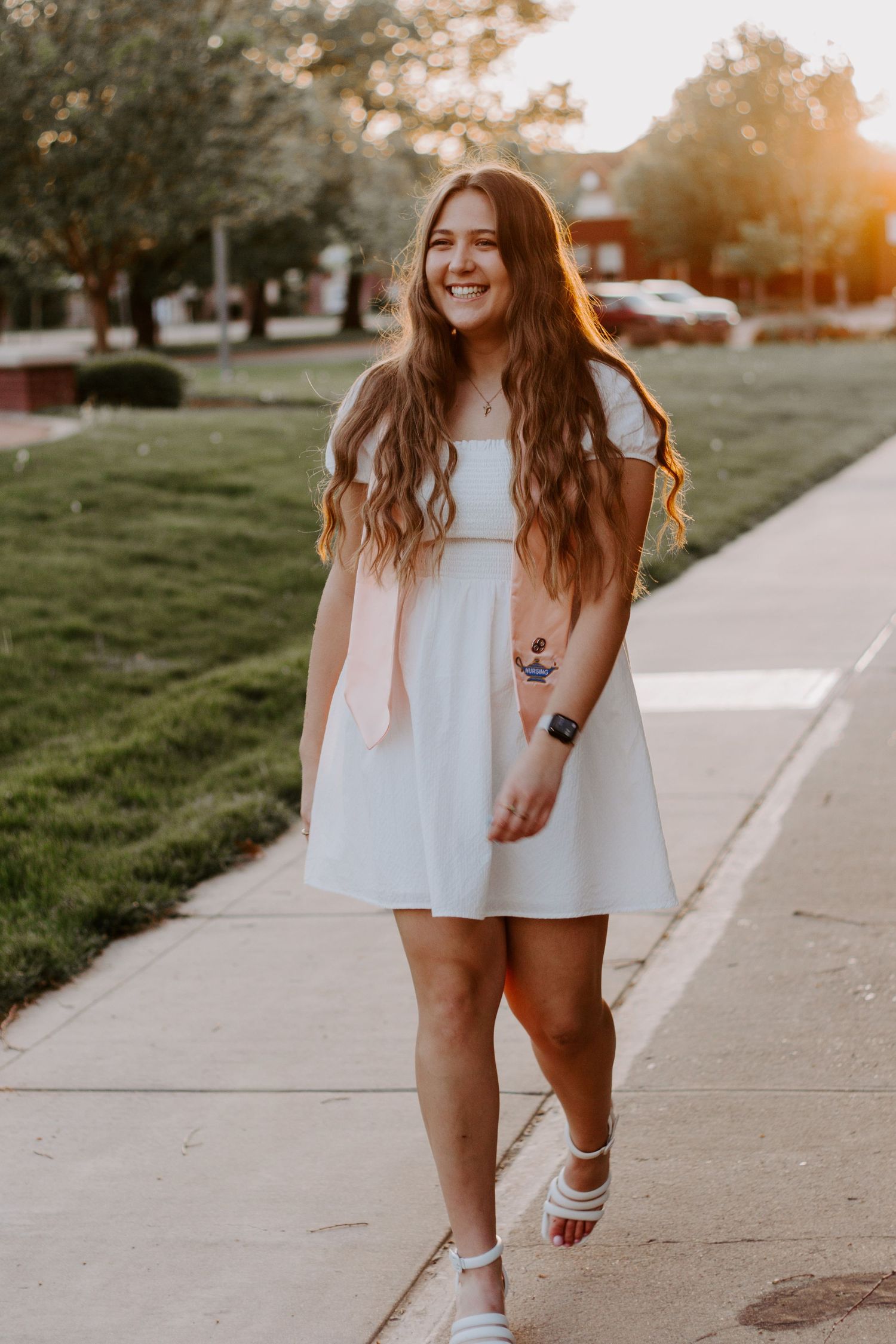 A smiling young woman with long wavy hair walks along a sunlit sidewalk in a white dress and white sandals.