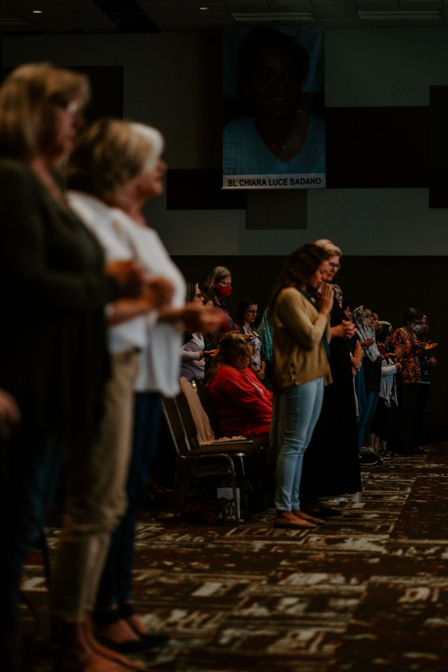 Attendees stand with hands folded in a dim conference hall beneath a large portrait banner during a faith gathering.