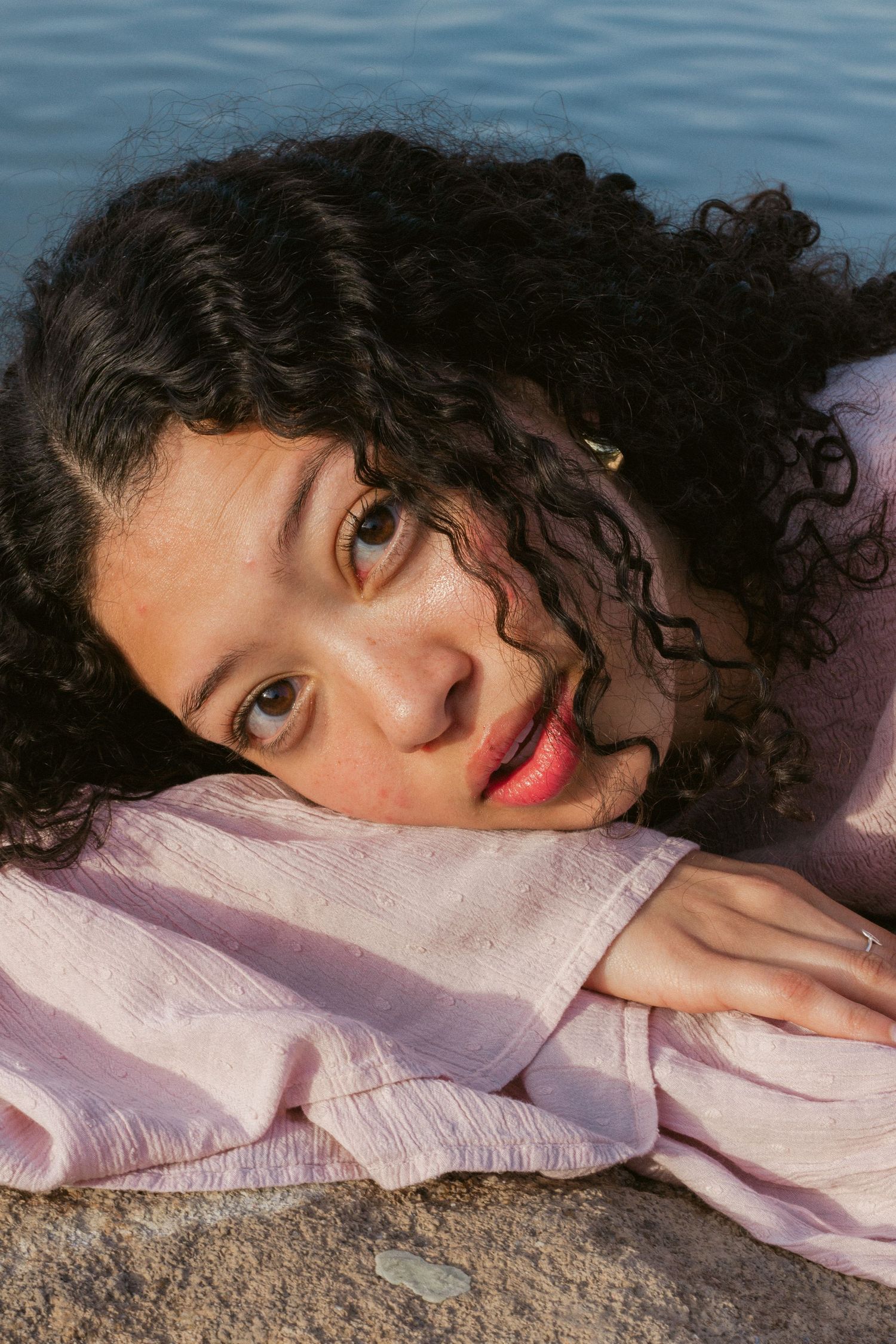 A young woman with dark curly hair rests her head on her arm beside the water, gazing at the camera in soft golden light.