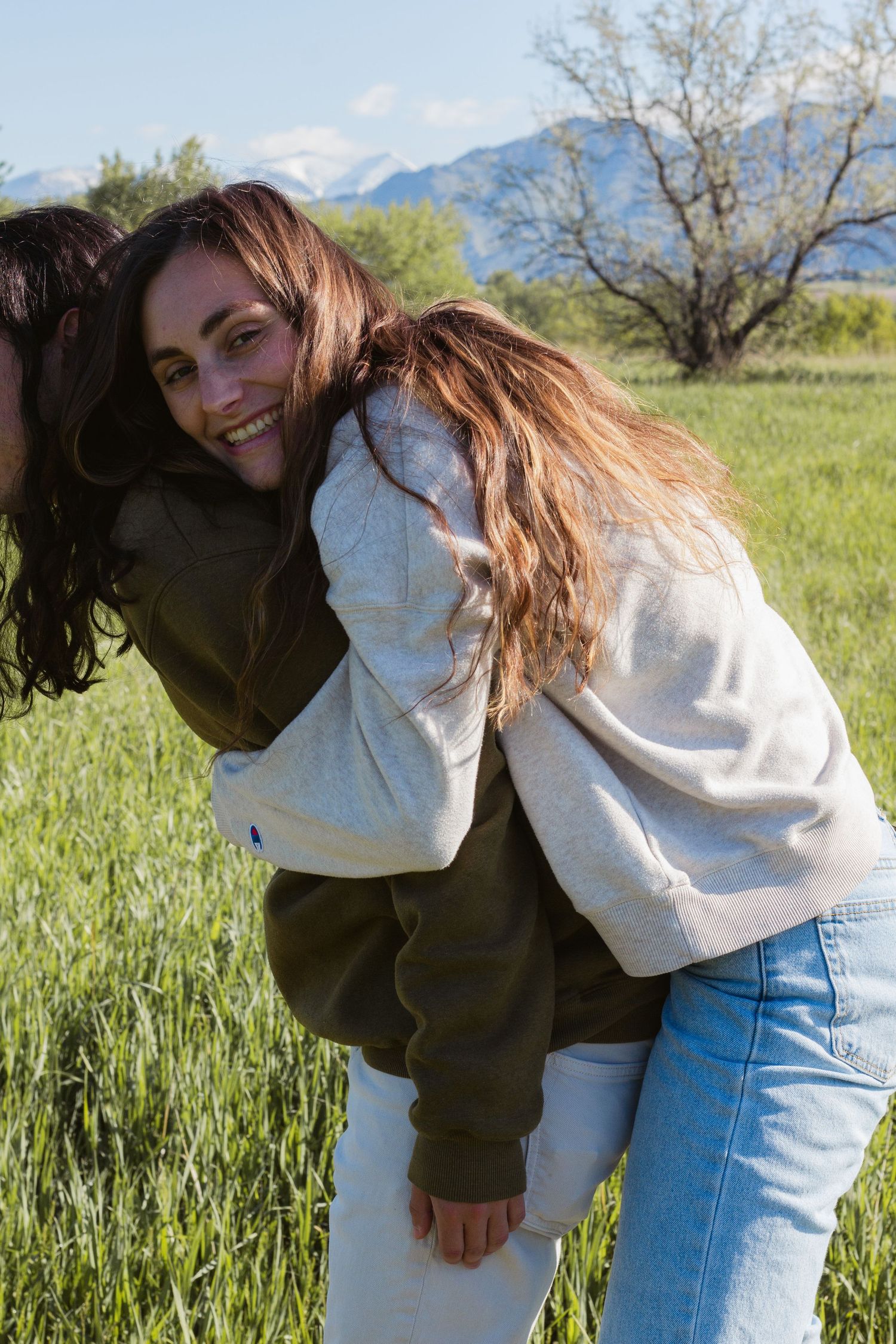 Two women laugh in a green mountain meadow as one gives the other a playful piggyback ride on a sunny day.