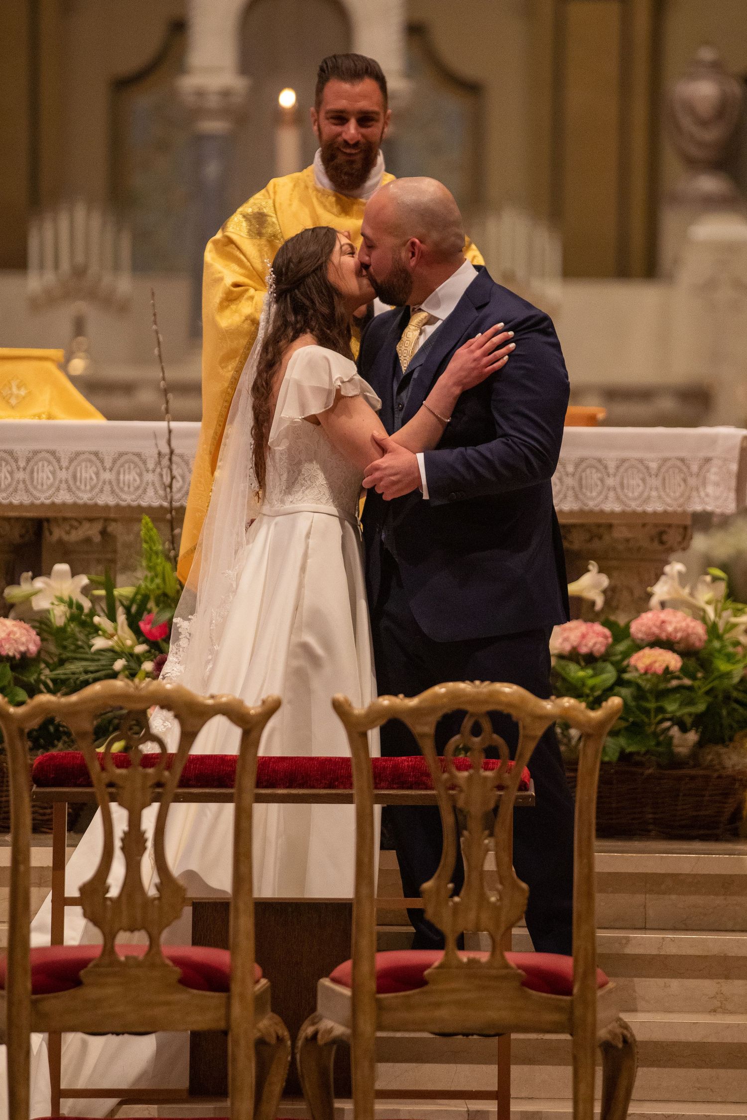 A bride in a white dress and a groom in a navy suit share a kiss at the church altar while a smiling priest in gold vestments looks on.
