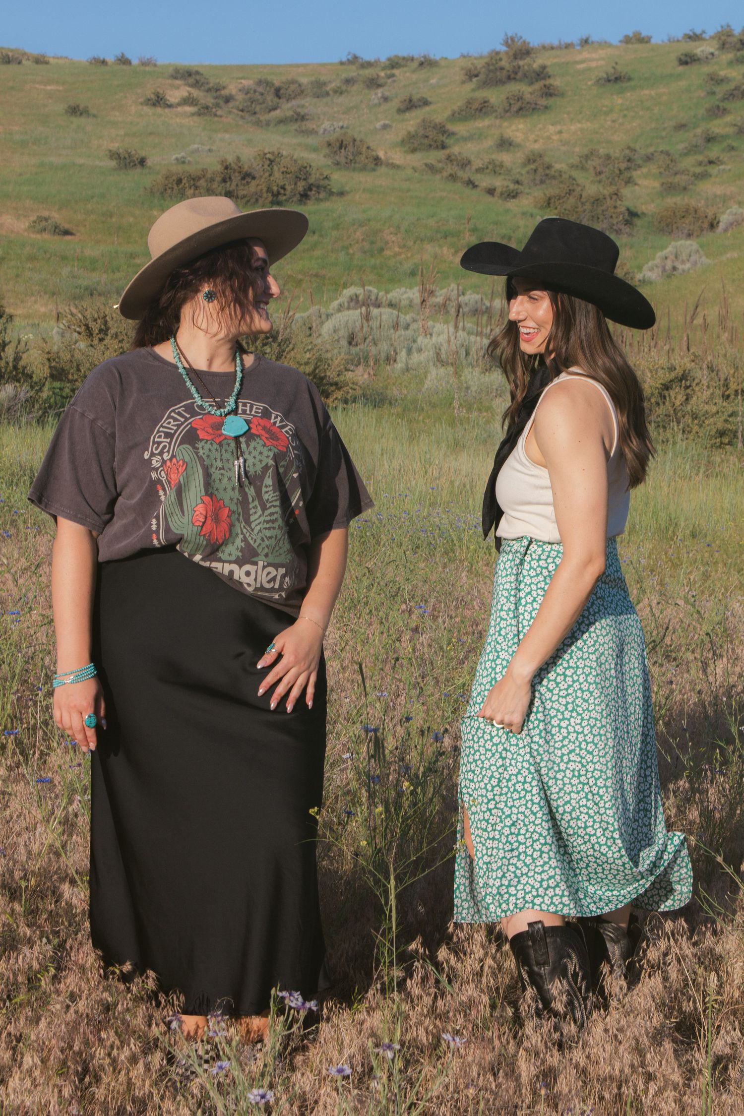 Two women in wide-brim hats stand facing each other and smiling in a sunny grassy hillside field.