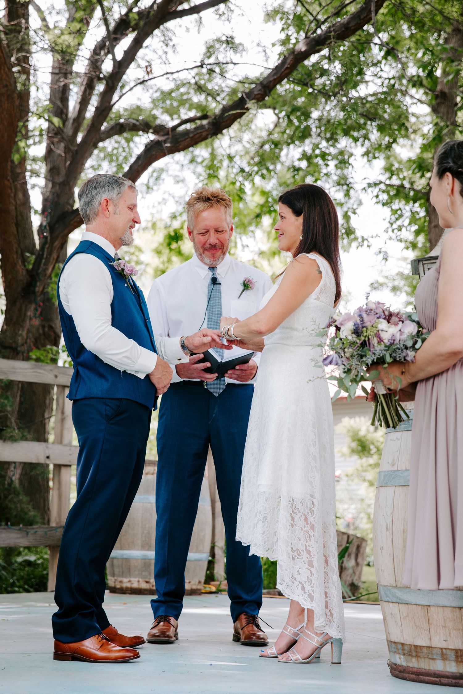 A bride in a white lace dress and a groom in a navy vest exchange vows with an officiant at an outdoor wedding ceremony.