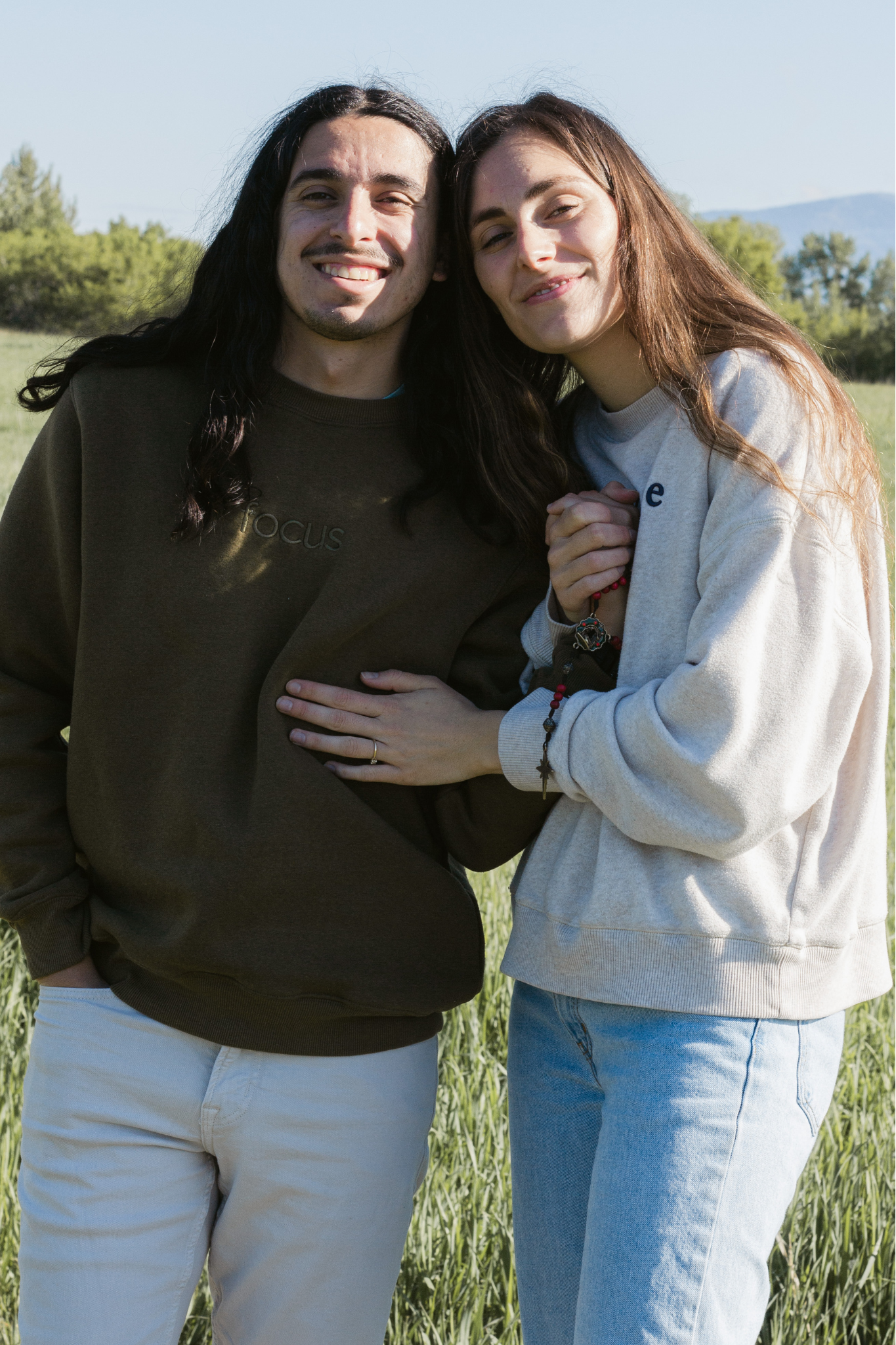 Smiling young couple embraces in a sunny green field, the long-haired man in an olive sweatshirt and the woman in a gray sweatshirt.