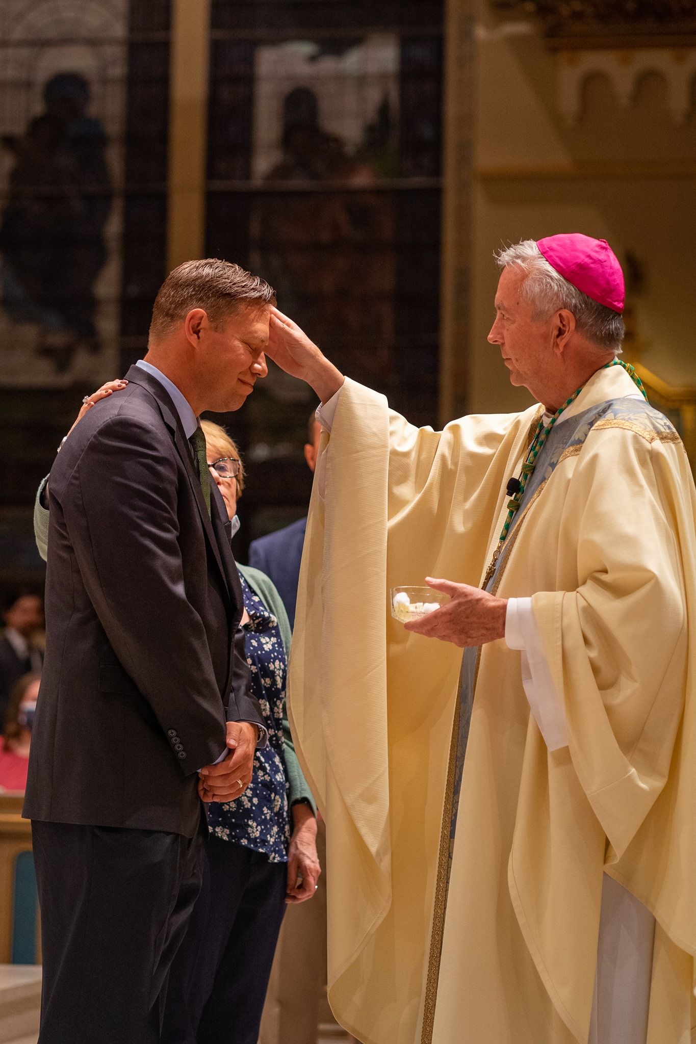 A bishop in a pink zucchetto and cream vestments places his hand on a man's forehead as a woman stands beside him during a church ceremony.
