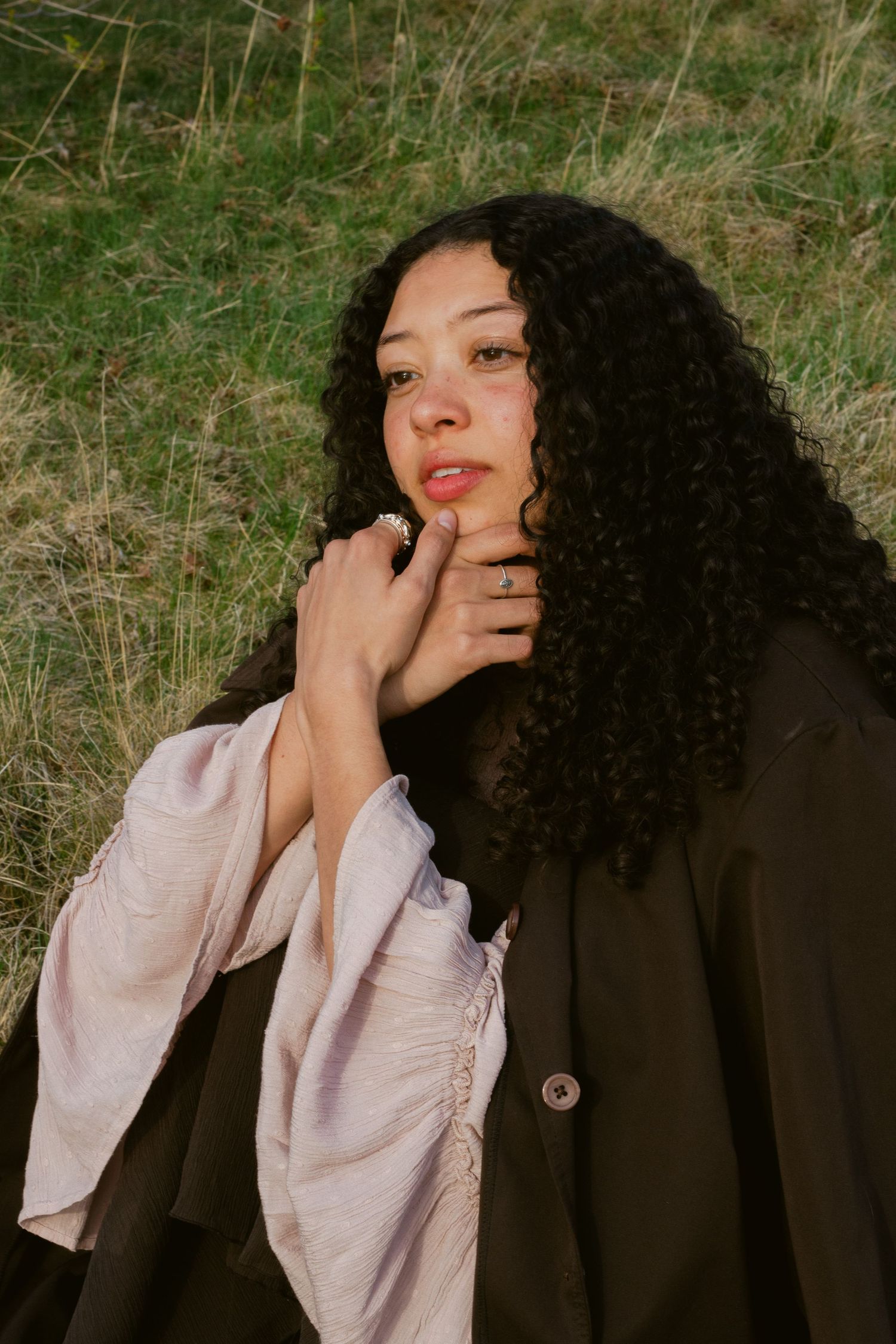 A young woman with long curly black hair sits on grass in a brown coat and pink flowing sleeves, hand raised near her chin.
