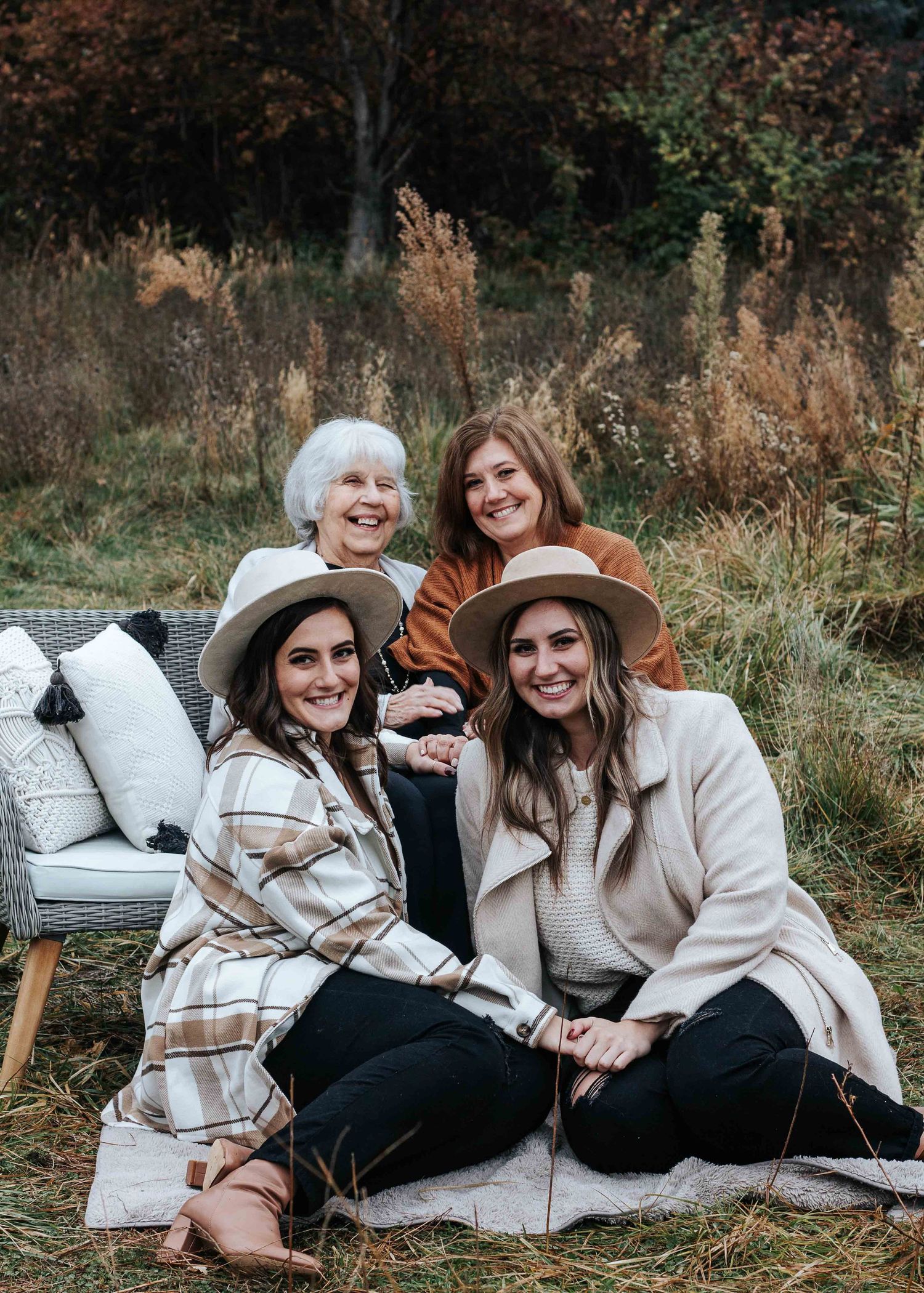 Four smiling women across generations pose together in an autumn field, two wearing wide-brim hats and cozy neutral layers.