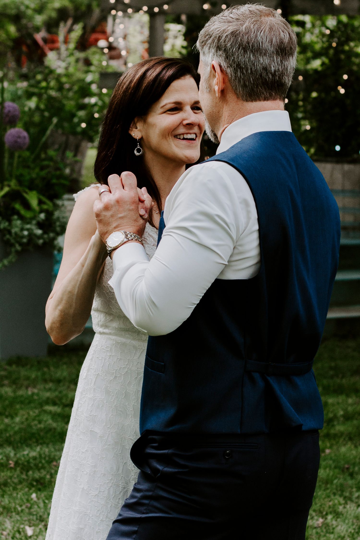 A bride in a white lace dress and a groom in a navy vest share a smiling dance in a garden with string lights.