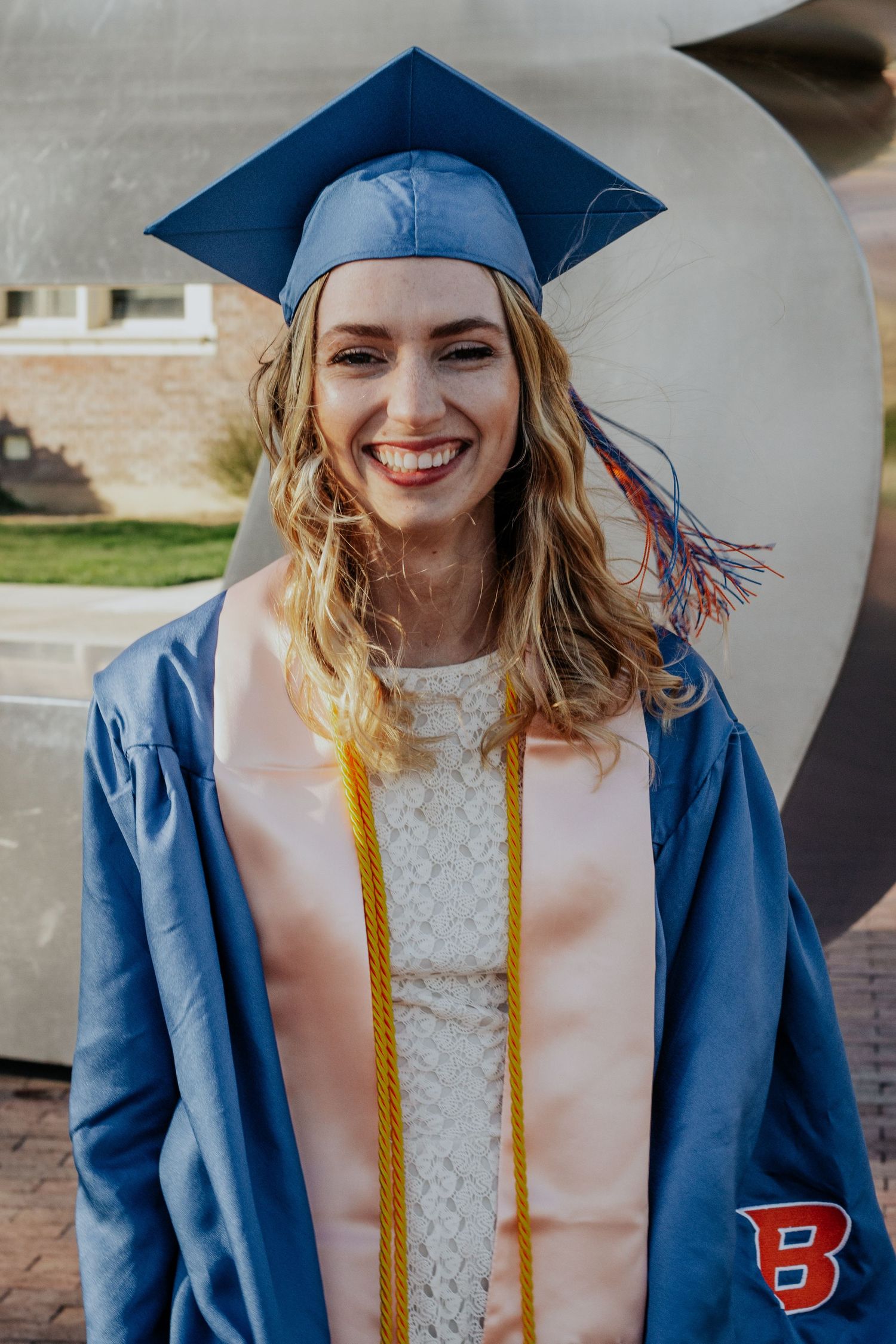 Smiling graduate with wavy hair wears a blue cap and gown, a peach stole and gold cords, with a Boise State B on the sleeve.
