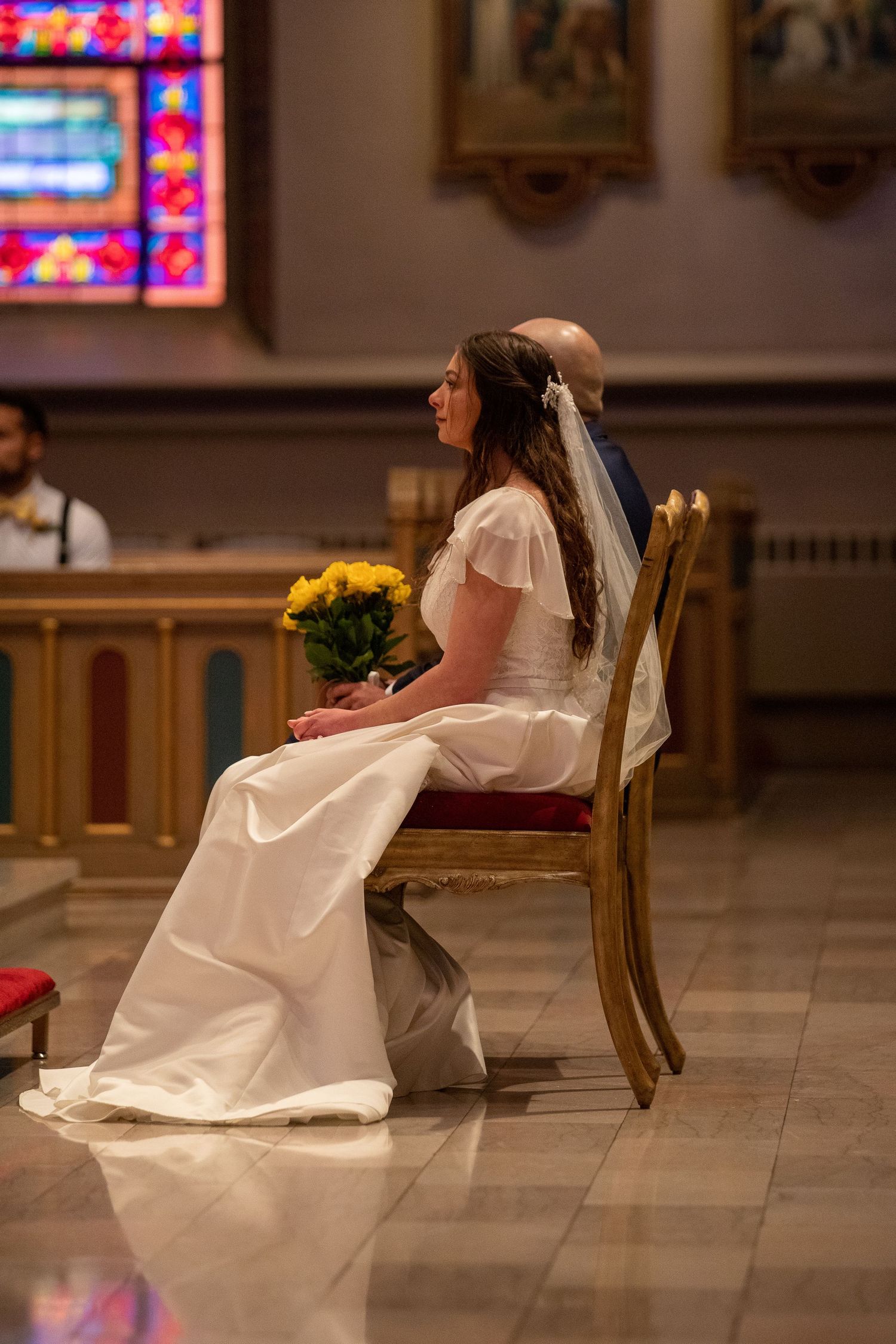 A bride in a white gown and veil sits beside her groom holding yellow flowers during a church wedding ceremony.