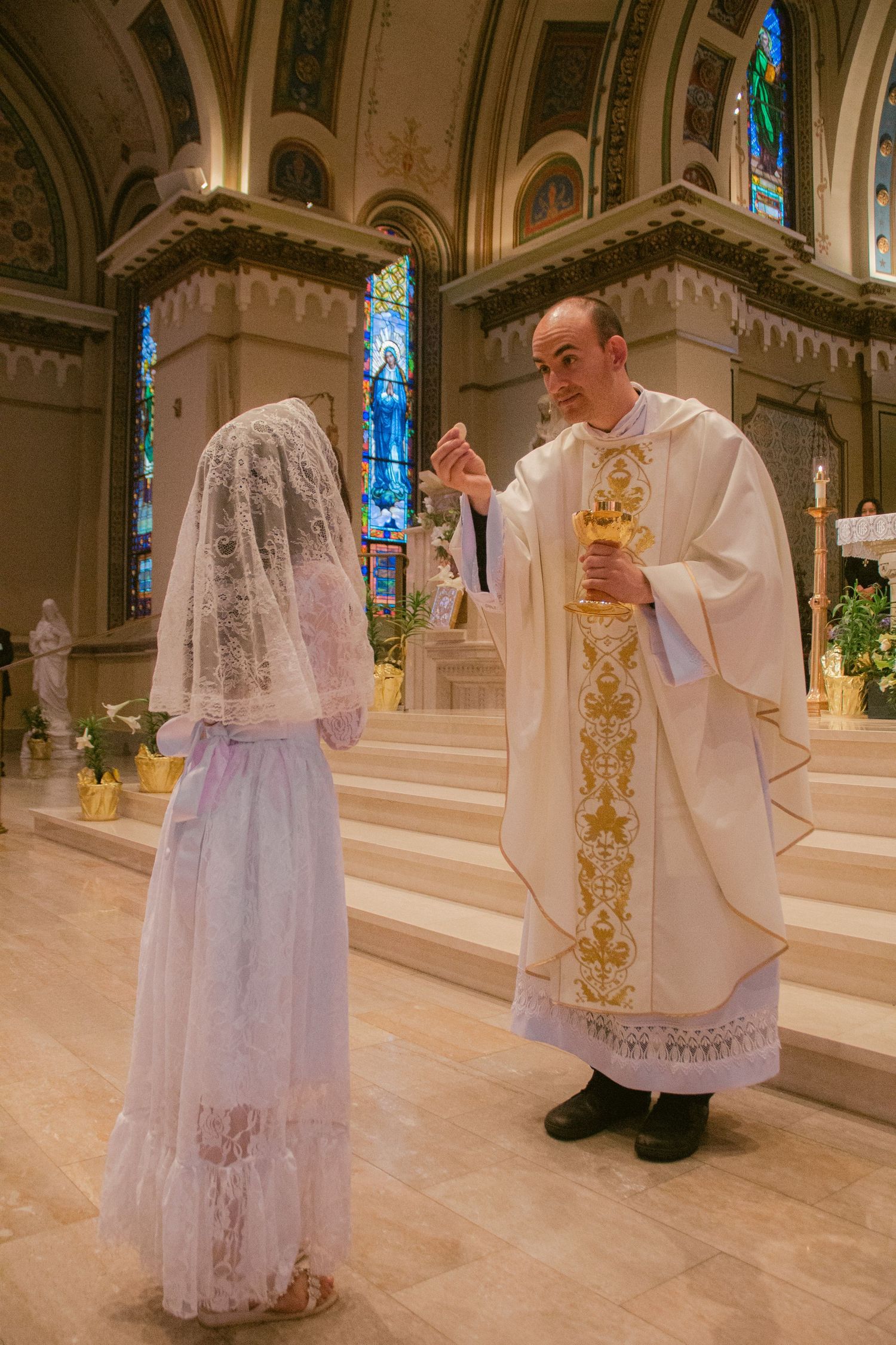 A priest in white and gold vestments raises a chalice before a girl in a lace veil and white dress at First Communion.