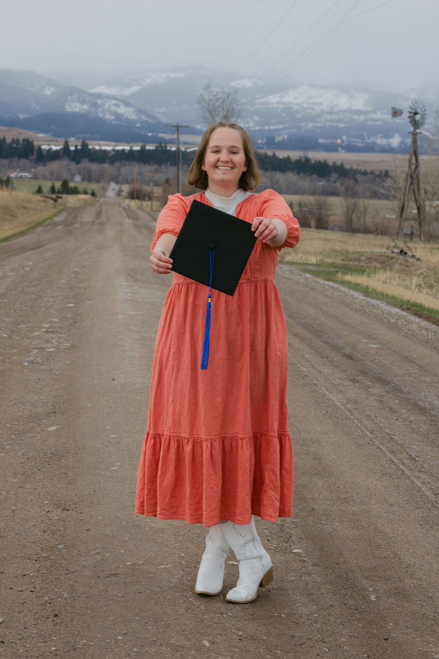 Smiling graduate in a coral dress and white boots holds out her black cap on a gravel road with snowy mountains behind.
