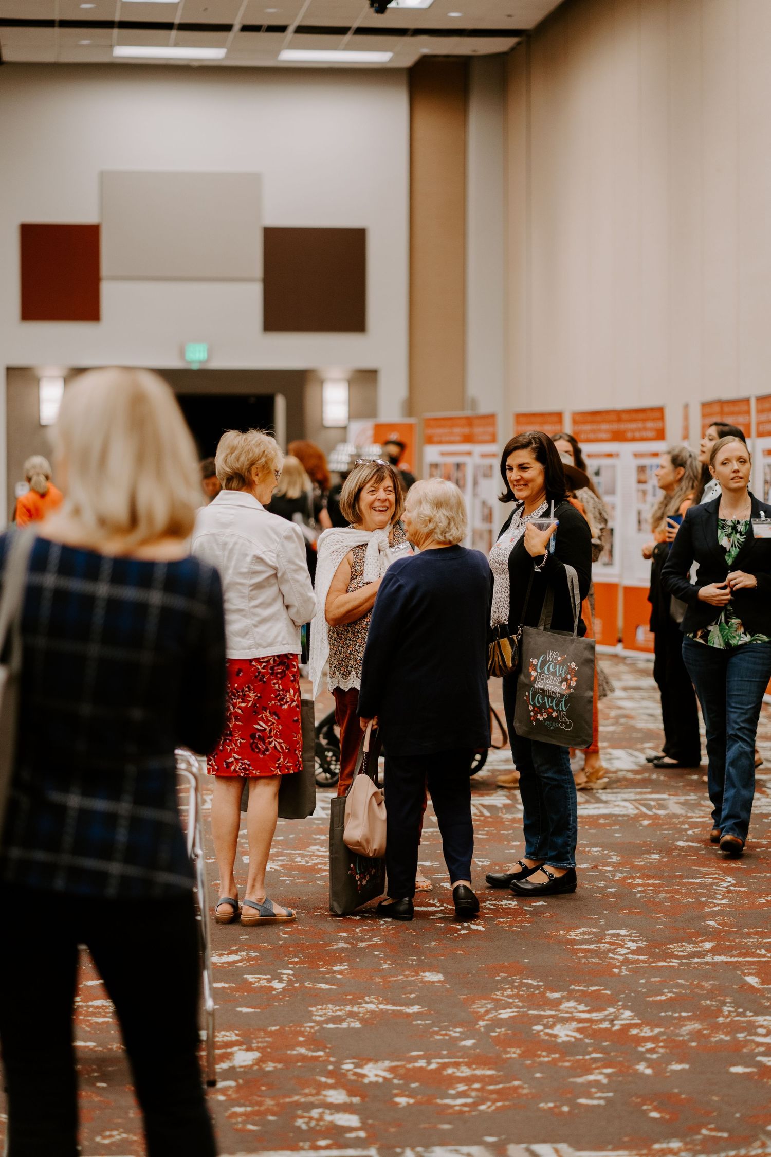 Women chat and network in a bright convention hall with informational display boards lining the wall behind them.
