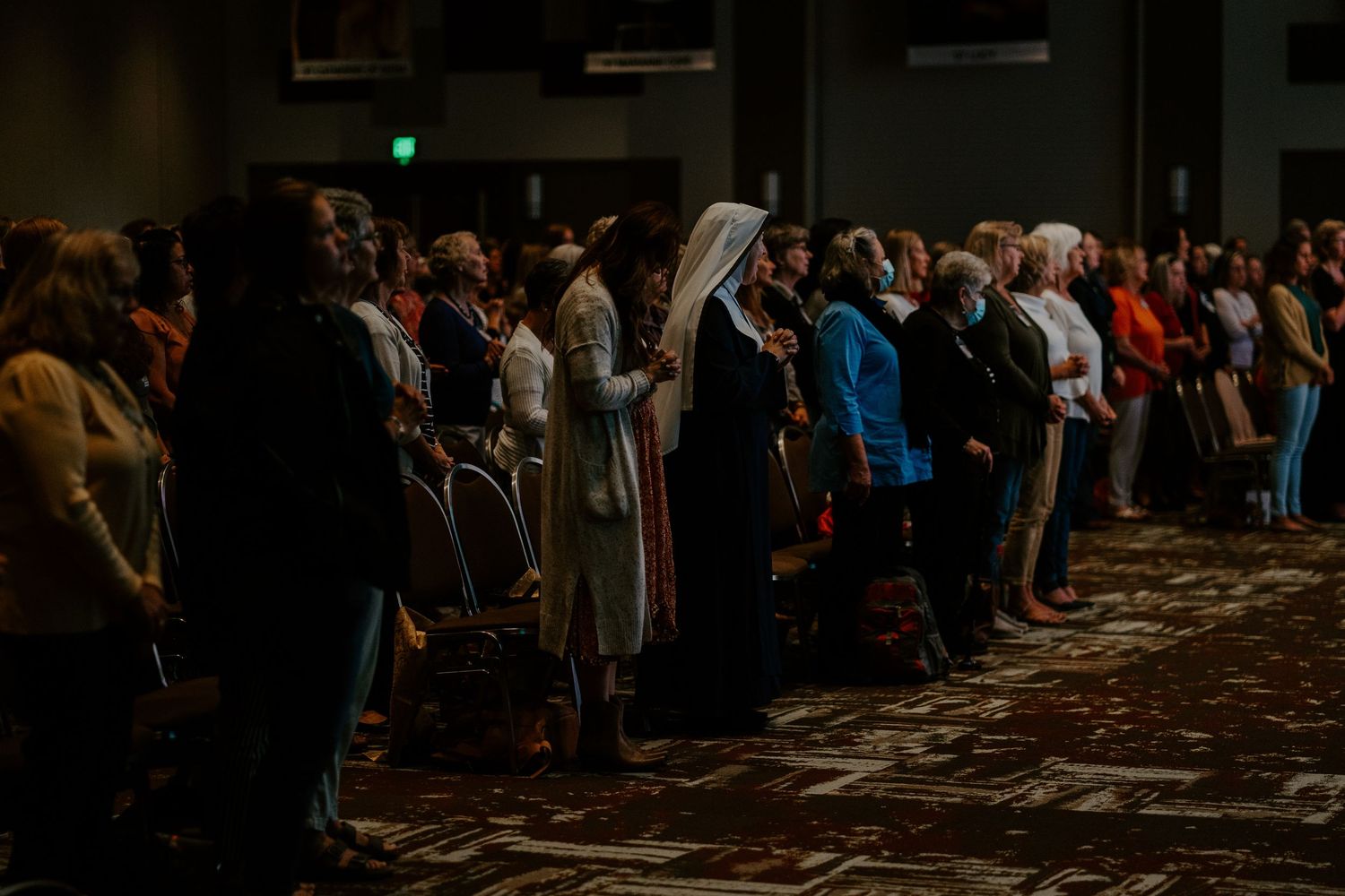 A large crowd stands together in a dimly lit conference hall, including women in religious habits, during a ceremony.