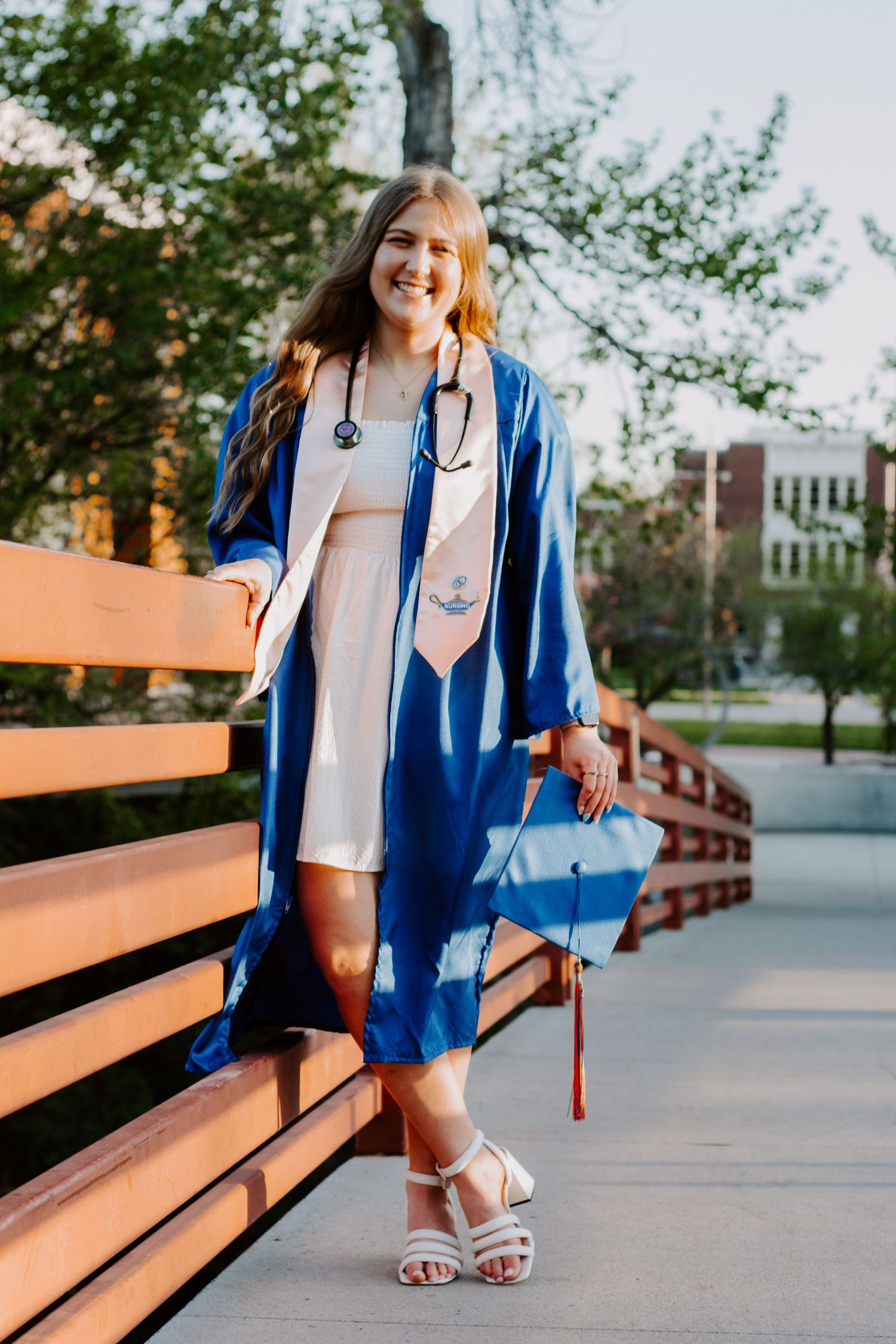 A smiling graduate in a blue gown and stethoscope poses on a wooden bridge holding her decorated graduation cap.