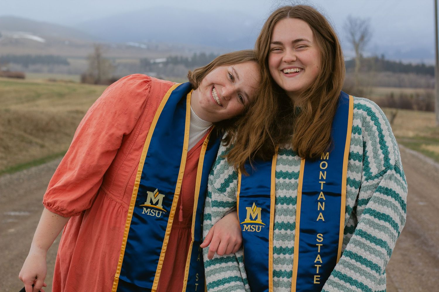Two laughing Montana State University graduates in MSU stoles embrace on a country road with rolling hills behind them.