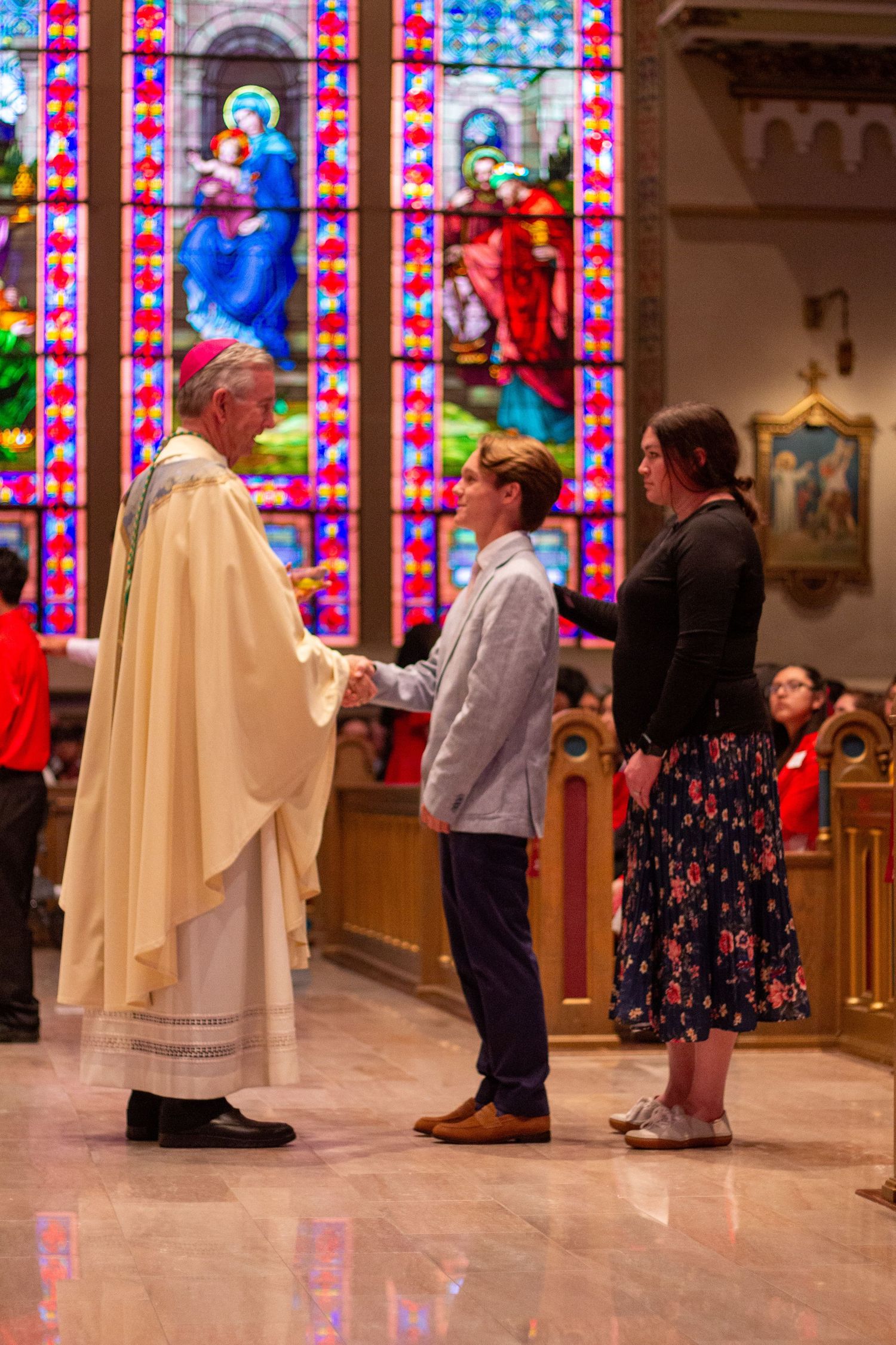 A priest in cream vestments shakes hands with a boy as a woman stands beside him in front of stained glass in a Catholic church.