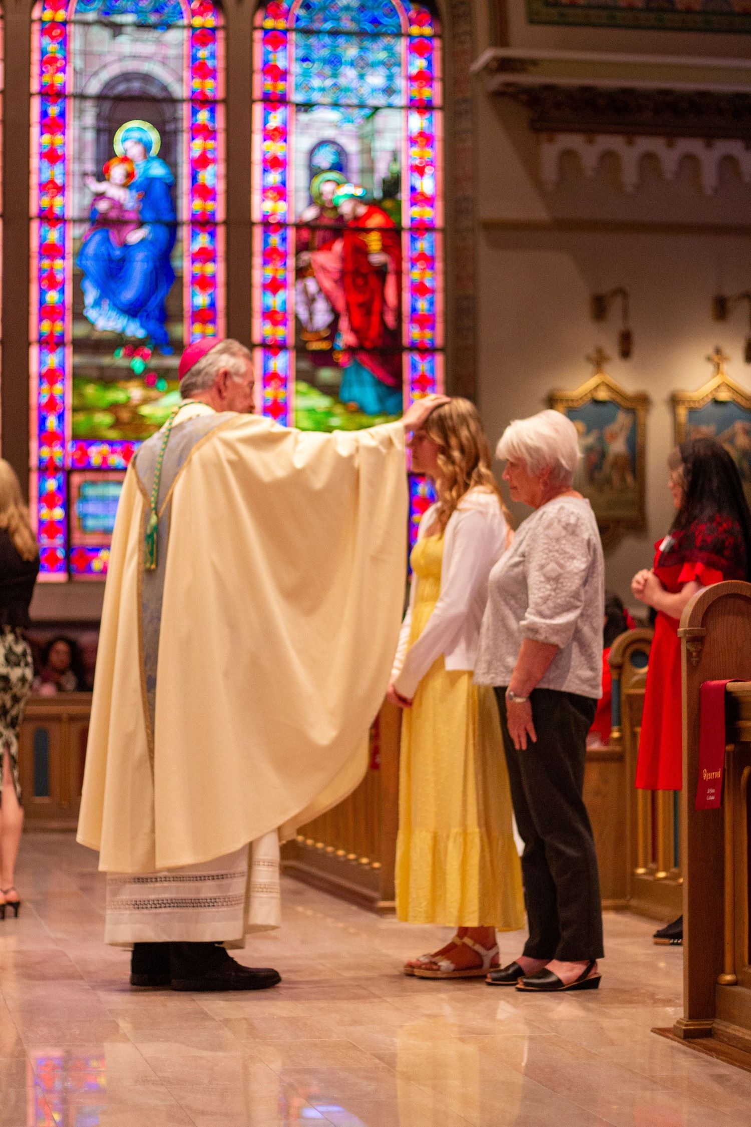 A priest in cream vestments extends his hand in blessing over two women standing before the pews in a church with stained glass windows.